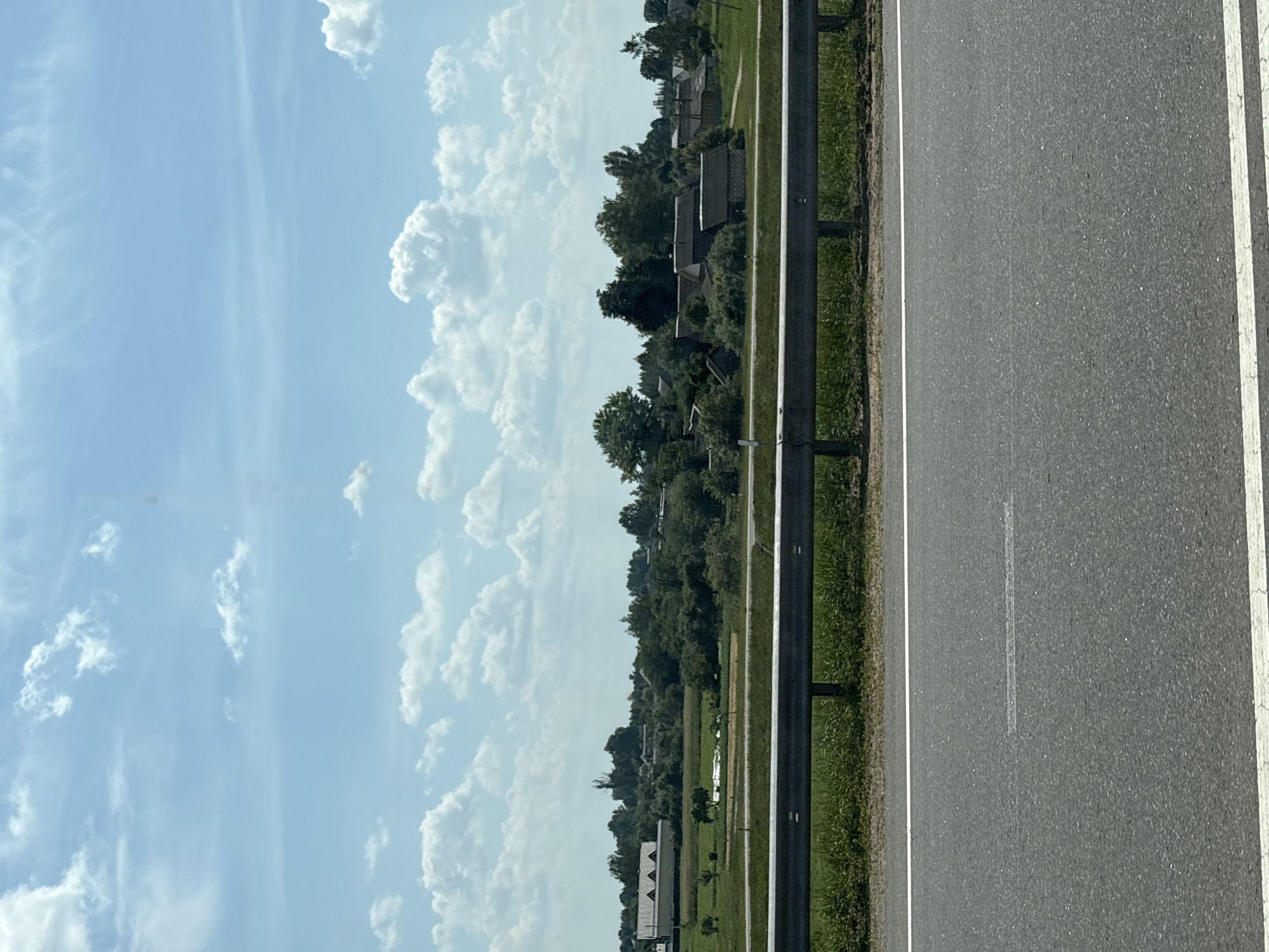 The image depicts a rural landscape with a clear blue sky filled with scattered white clouds. In the foreground, there is a section of asphalt road with white lane markings, bordered by a metal guardrail and grassy verges. Beyond the guardrail, a green field stretches towards a cluster of buildings, suggesting a small village or hamlet. The buildings appear to be residential, with pitched roofs and varying architectural styles, surrounded by trees. The overall impression is that of a tranquil, sunlit day in a countryside setting, possibly near Мастоцкі сельскі Савет, Belarus. No people or animals are visible in the frame.