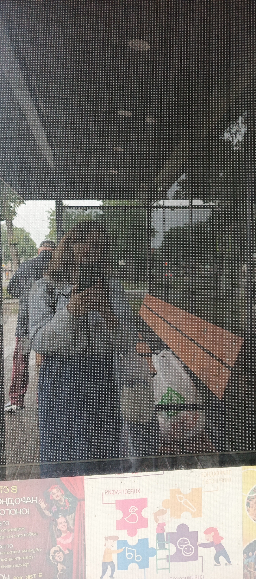 The image appears to be a reflection on a screen or window, showing a street scene. In the foreground, a woman is visible, taking a picture with her phone. She is wearing a grey jacket and dark pants. Behind her, other people and a white plastic bag are also visible. The background shows trees and a cloudy sky, suggesting an outdoor setting in a city. There are posters or advertisements visible at the bottom of the frame, one of which seems to have text in Cyrillic.