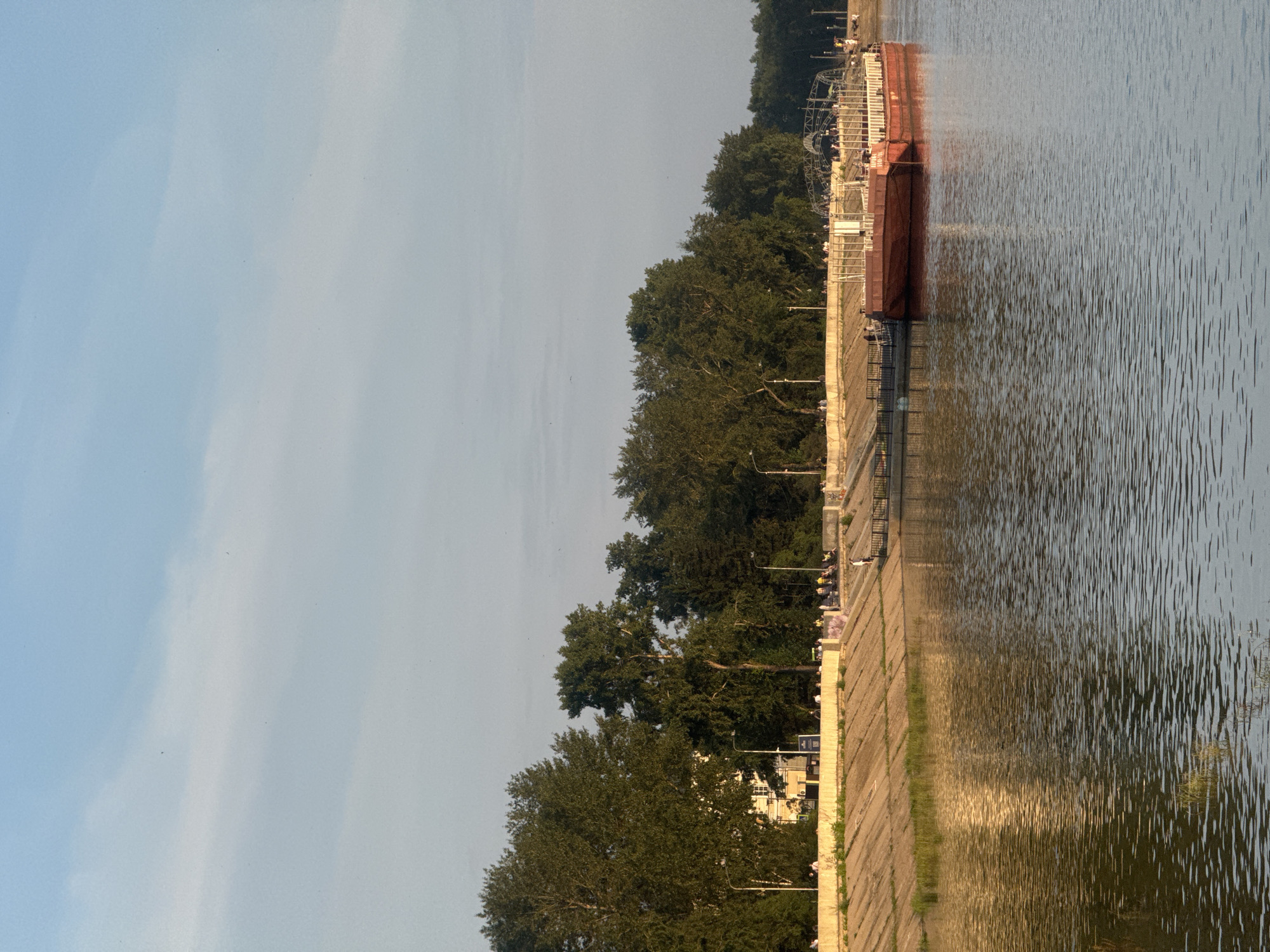 A wide river flows from left to right in the foreground, reflecting the golden light of a setting sun. On the right bank, a red barge with a white superstructure is moored to a pier extending into the water. Beyond the barge, a concrete embankment rises, topped with a walkway lined with trees. People are visible walking and sitting on the embankment and walkway. The sky is a pale blue with thin wisps of clouds. The scene is likely the Angara River embankment in Irkutsk, Russia, during the evening.