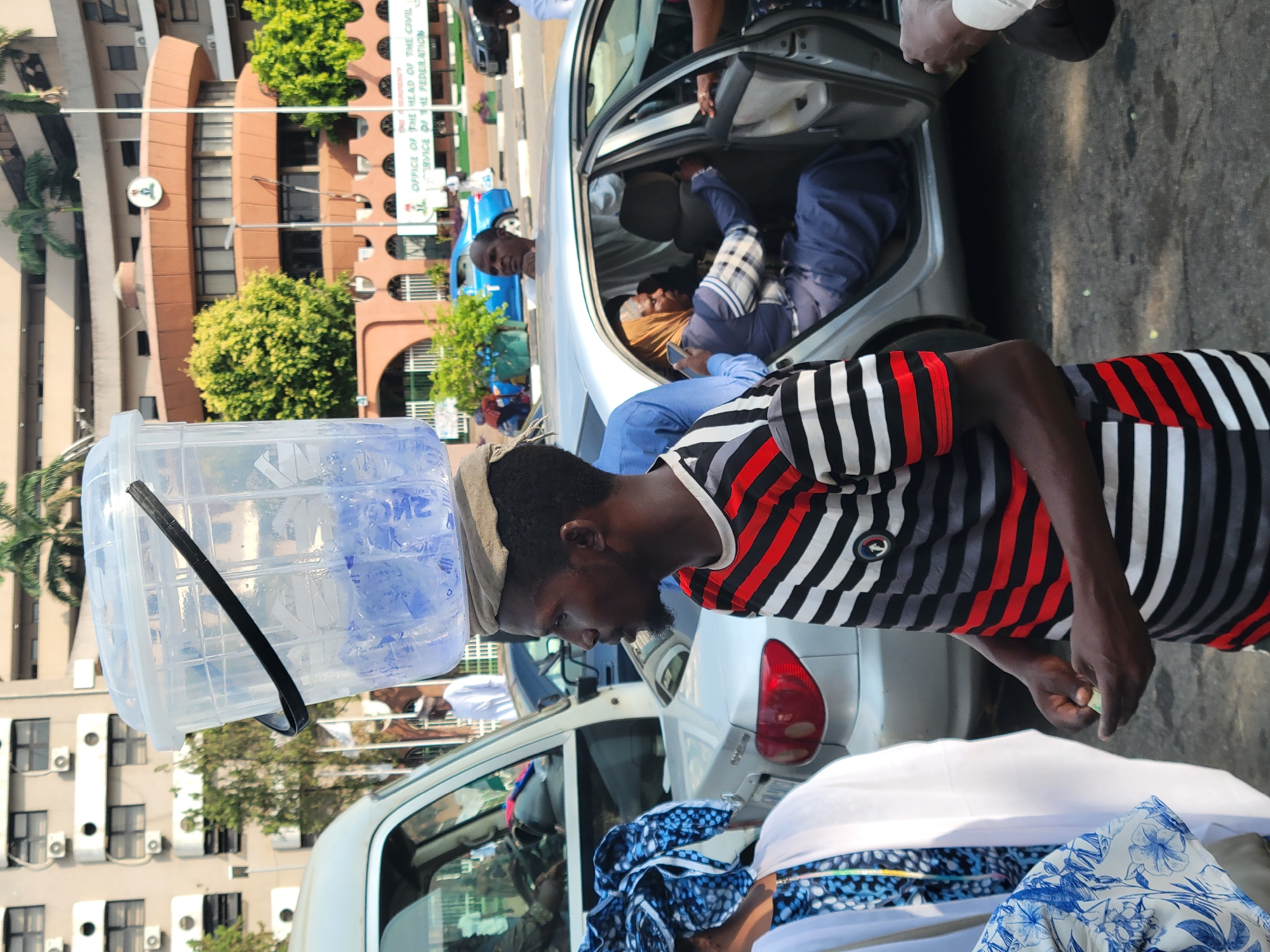The image depicts a scene in Abuja, Nigeria, in front of the "Office of the Head of the Civil Service of the Federation." A man wearing a striped shirt and a cap is standing with a large water container balanced on his head. Adjacent to him is a silver car with its door open, and people are visible inside. Other individuals are nearby, and their actions and interactions are unclear. The surroundings consist of buildings, trees, and other vehicles. The overall setting appears to be a busy public area.
