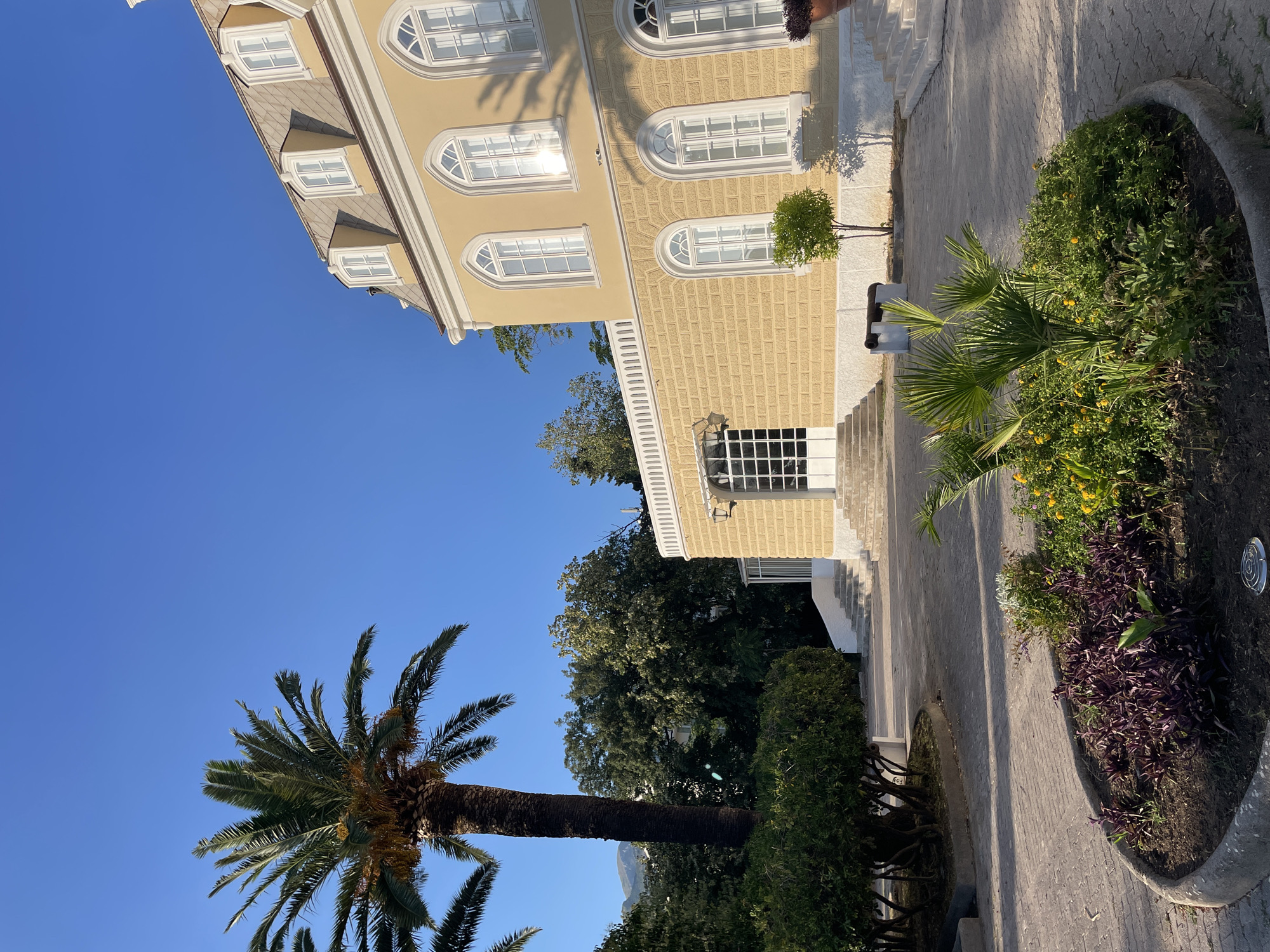 The image shows a sunny day in Bar, Montenegro, with a clear blue sky. The scene features a light yellow building with arched windows and a tiled roof, likely a historical structure. In front of the building, a paved pathway leads to a garden area with a variety of plants, including a palm tree and some purple flowering bushes. The sunlight casts shadows, indicating it's likely morning or late afternoon. The building's architecture suggests a European style, possibly from the 19th or early 20th century. The overall atmosphere is peaceful and serene, with no people visible in the frame. The image is oriented vertically, with the sky occupying the top left portion and the building and garden filling the rest.