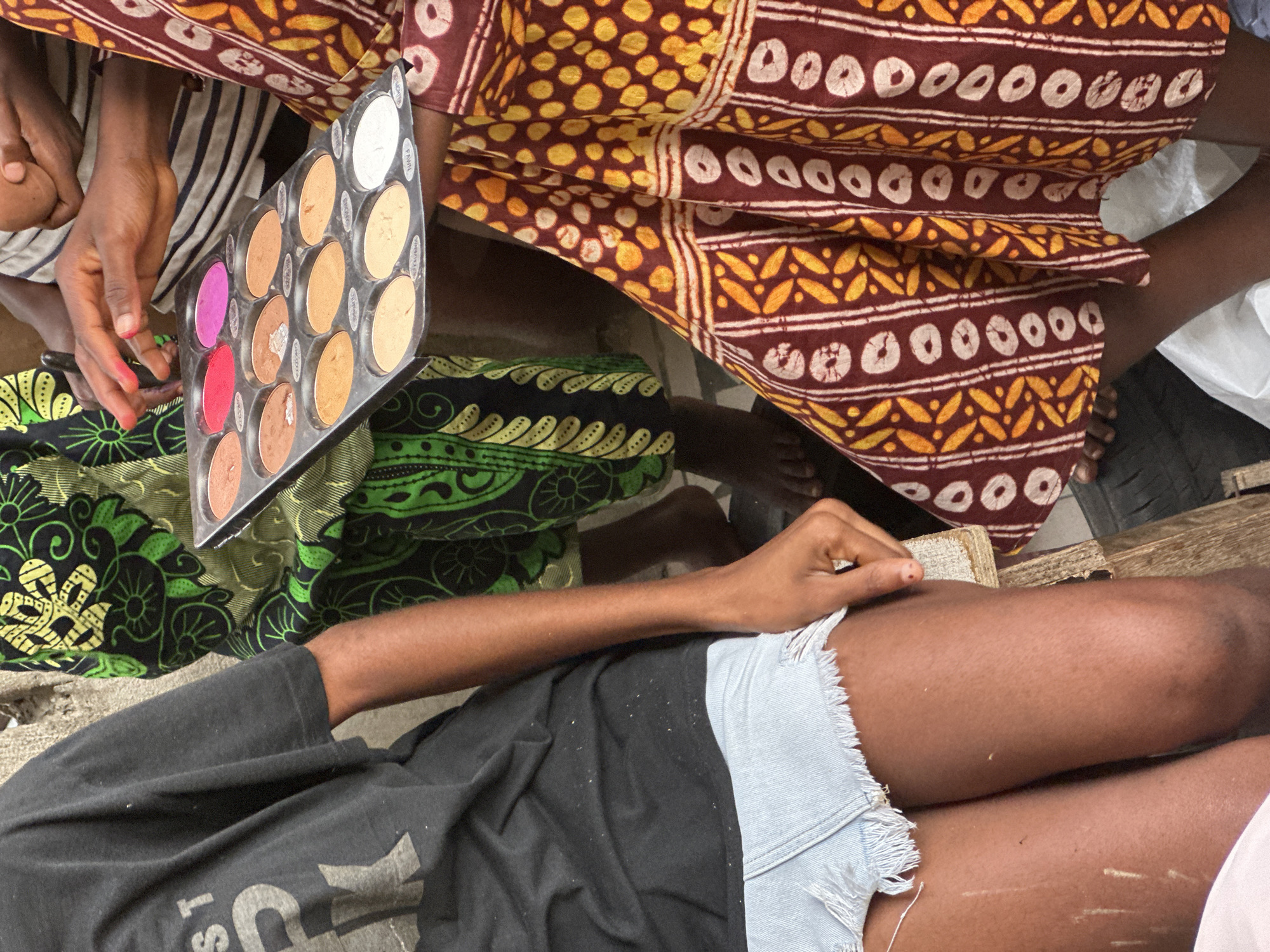 In Badagry, Nigeria, a close-up shot focuses on a group of individuals. One person, wearing a black t-shirt and light blue denim shorts, is seated on the left. To the right, another person in a vibrant, patterned garment is holding a makeup palette containing various shades of foundation and pink eyeshadow. A third person, partially visible in the background, is wearing a black and white striped top. The scene suggests a makeup application or demonstration is taking place outdoors. The lighting is natural and diffused.