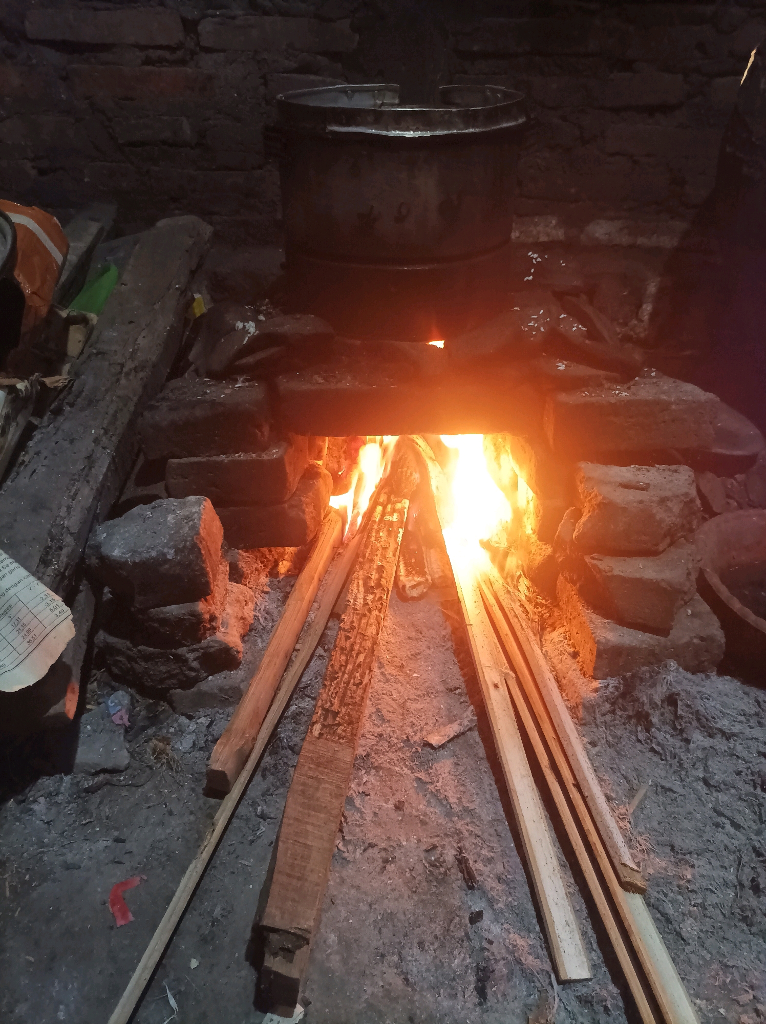 The image depicts a traditional wood-burning stove with a pot placed on top. The stove is constructed from bricks and filled with burning wood, producing a bright orange and yellow flame that illuminates the scene. Several pieces of firewood are visible, both inside the firebox and arranged around it. A large, dark-colored pot is situated over the opening of the stove, suggesting that food is being cooked. The background shows a brick wall. Scattered on the ground are ashes, some debris, and a crumpled piece of paper with text and numbers. The overall environment appears to be an outdoor or semi-enclosed cooking area, possibly in a rural setting. The location is identified as Canga'an, Indonesia. There are no people visible in the image.