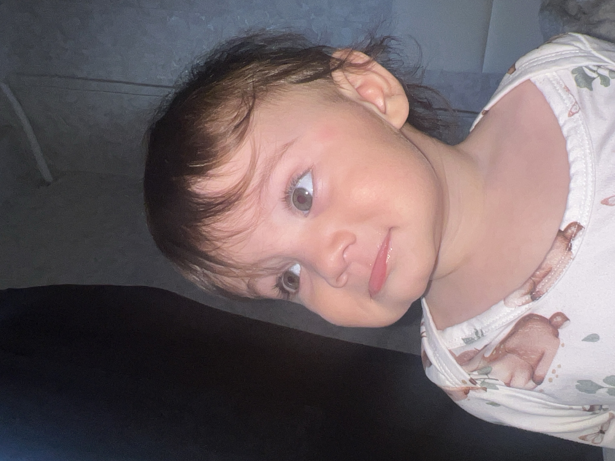 The image is a close-up portrait of a young child, likely a baby or toddler, captured with a tilted perspective. The child's head is angled, and they are looking directly into the camera with a wide-eyed, engaging expression.
The child has fair skin, dark brown hair with lighter brown highlights, and striking light-colored eyes, appearing to be green or hazel. A slight, perhaps curious or playful, smile is present, with the tip of the tongue visible between slightly parted lips. The child is wearing a light-colored, possibly white or cream, garment, which appears to be a top or bodysuit. The fabric features a repeating pattern of small, light brown animals, possibly bears or squirrels, interspersed with green leaf designs, resembling ginkgo leaves or similar foliage.
The background is dark and indistinct, suggesting an indoor setting. Areas of dark gray and black dominate, with some subtle textures and lines visible, indicating furniture such as a couch or bed, and possibly a wall. The lighting appears to be artificial, illuminating the child's face against the darker surroundings.
No specific activity is taking place other than the child engaging with the camera. The child's expression conveys curiosity and perhaps a hint of playfulness. There are no discernible cues within the image to indicate the specific time of day, weather conditions, or particular location within Elena, Bulgaria. No text is visible.