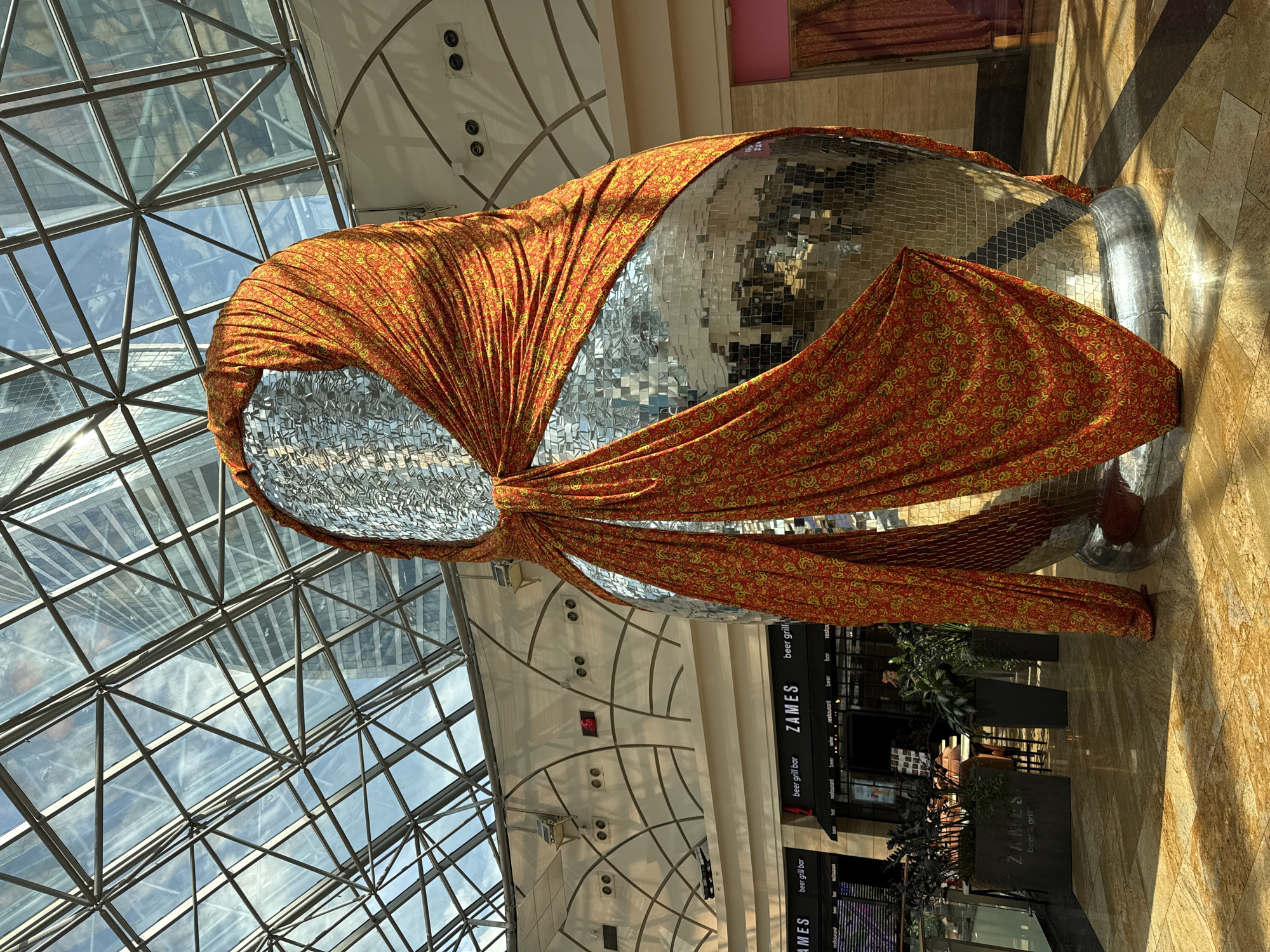 Here's a description of the image:
The image shows an indoor scene, presumably within a shopping mall or large commercial space. A large, disco ball-like structure, partially covered in a vibrant orange and yellow patterned fabric, is the focal point. The structure's surface consists of many small mirrored squares. The fabric appears draped over the top and sides of the object.
The location features a modern architectural design with a glass ceiling made of interconnected triangular panes, allowing natural light to flood the space. Below the structure, there's signage for "ZAMES," which appears to be a beer grill/restaurant. Some greenery in planters is also visible near the restaurant.
The image appears to be taken within the MEGA Khimki shopping mall, as this location features a similar architectural style and contains the ZAMES restaurant.