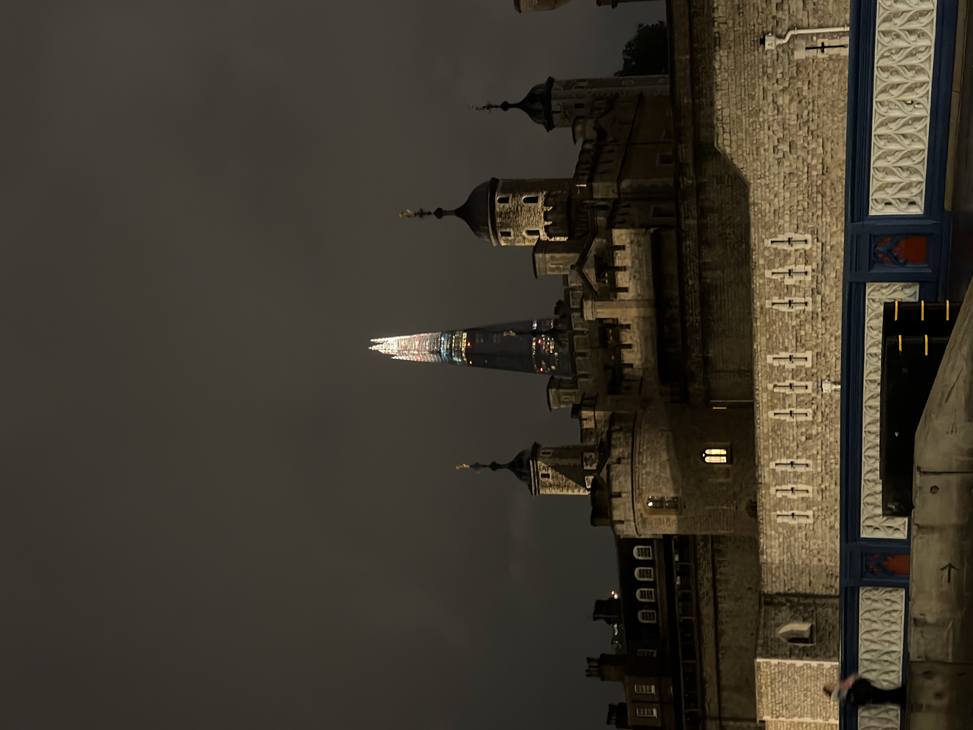 The Tower of London and The Shard are illuminated at night in London, United Kingdom. The Tower Bridge is partially visible in the foreground. The sky is dark and overcast.