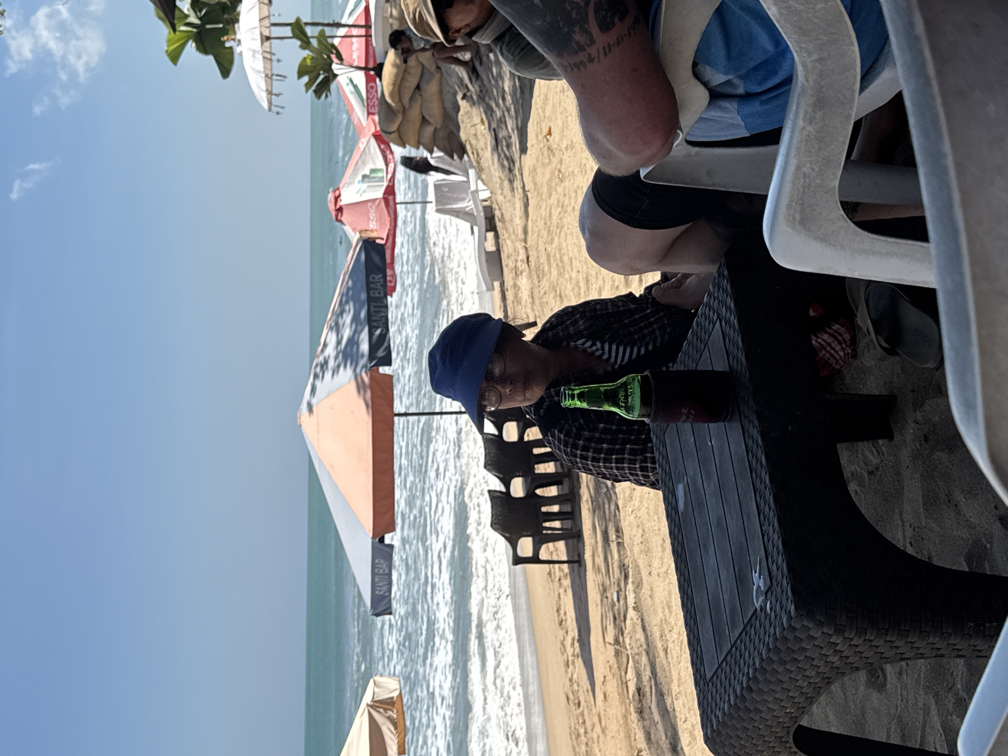 The image depicts a beach scene in Kuta, Indonesia, on a clear day. In the foreground, a black plastic table is set on the sand. A woman wearing a blue hat and a plaid shirt sits at the table, facing left. A green glass bottle is positioned on the table in front of her. To the right of the woman, a person's legs are visible, wearing black shorts and blue and white striped shorts. In the background, the ocean with waves is visible, with several beach umbrellas providing shade. Behind the umbrellas, sandbags are stacked, and other people are seated in chairs or walking on the beach. The overall environment suggests a casual beachside establishment.