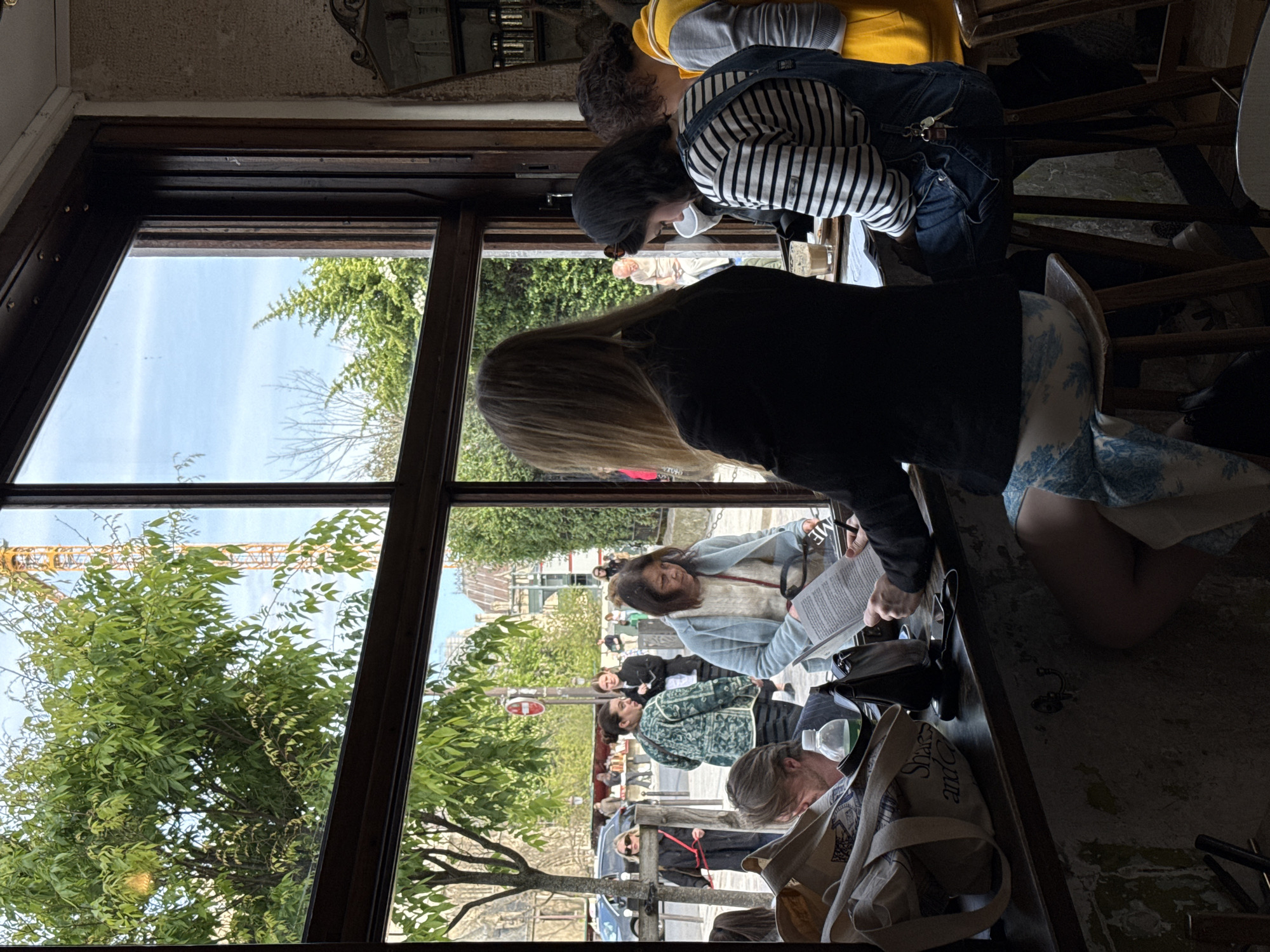 Inside a cafe, a group of people are seated at a window counter, reading and looking out at a Parisian street. The view through the window reveals trees, buildings, pedestrians, and traffic. A construction crane is visible in the distance. The cafe interior appears worn, with textured walls and vintage decor. The individuals are diverse, with various clothing styles and hairstyles. They seem engaged in personal activities, reading or observing the outside world. A variety of personal belongings, such as bags and purses, are scattered on the counter. The overall atmosphere is casual and relaxed.
