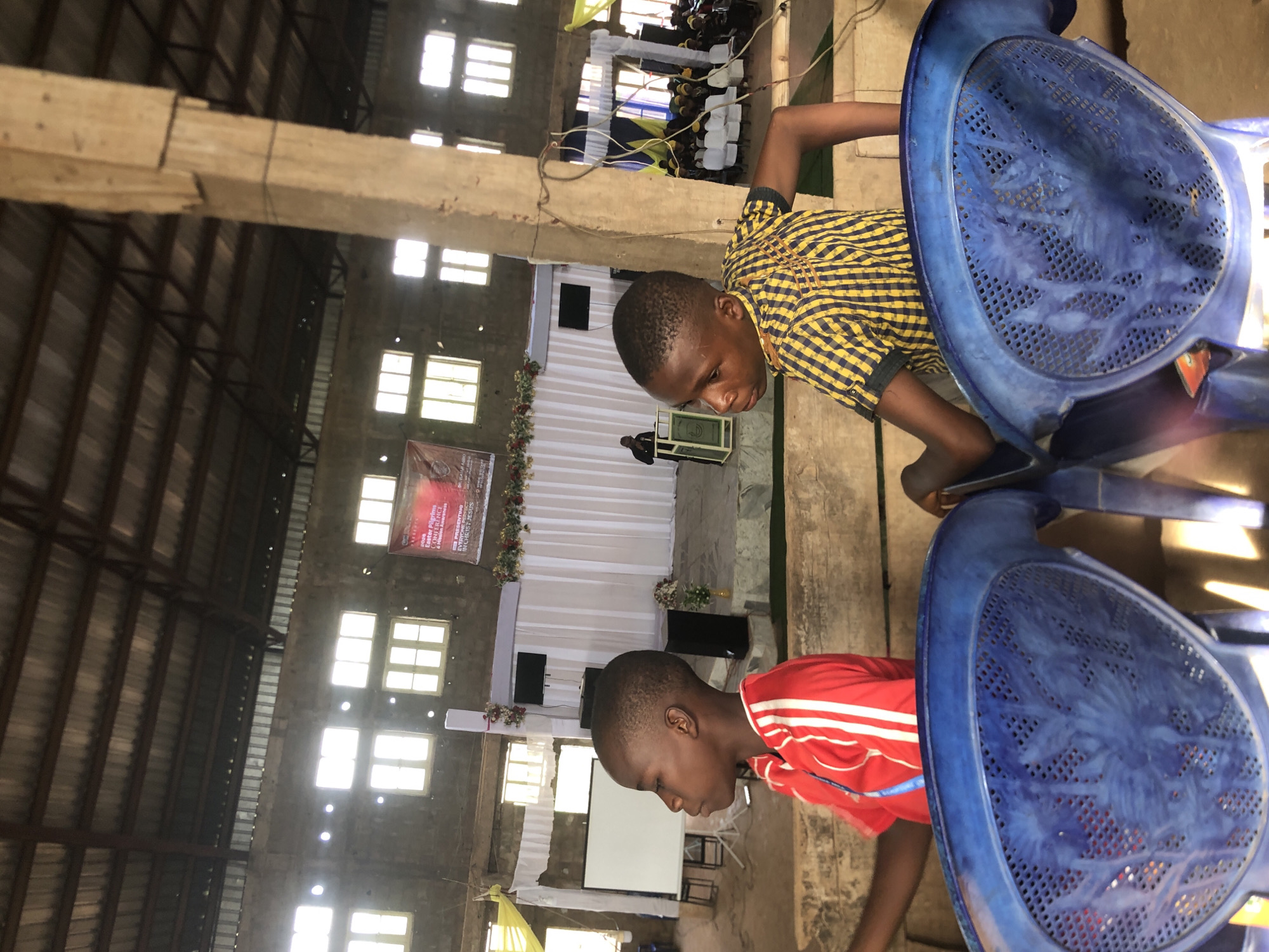 A medium, angled shot inside what appears to be a large hall or church in Nteje, Nigeria. Natural light streams through numerous windows, illuminating the interior. The hall has a high ceiling with exposed beams and a metallic roof. At the far end of the hall, a stage is decorated with white fabric, flowers, and a large banner. A man stands at the podium on the stage.
In the foreground, two young Black boys are positioned near rows of blue plastic chairs. One boy wears a yellow and black checkered shirt, and the other wears a red shirt with white stripes. They both appear to be looking downward, possibly observing something on the floor or ground. The blue chairs are stacked close to the camera, partly obscuring the view of the boys.