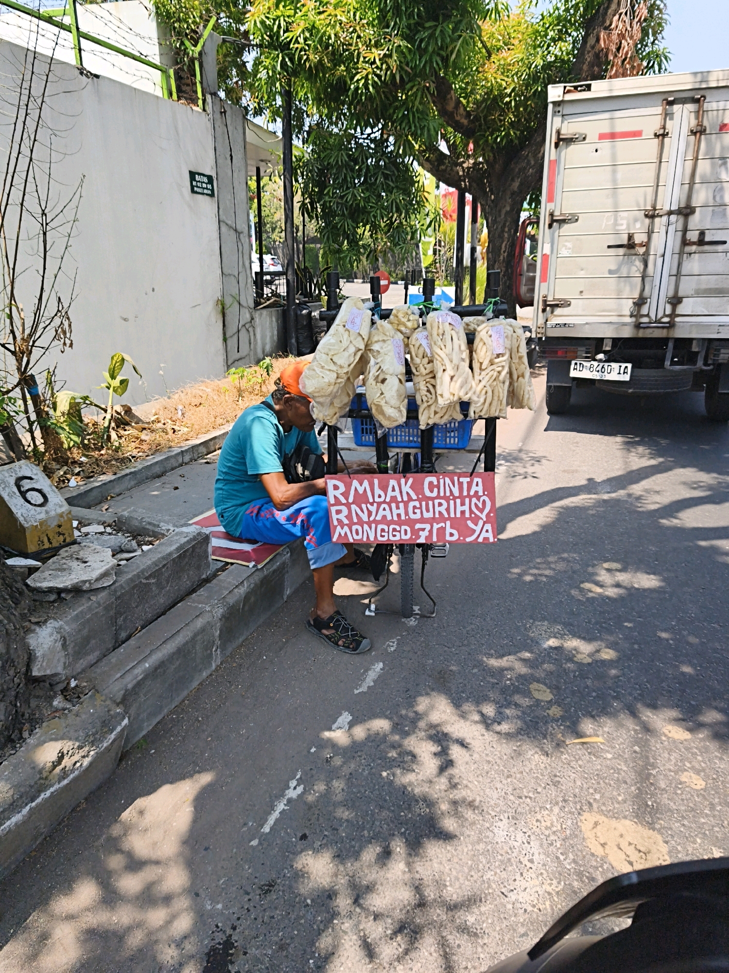 The image shows a man sitting on a red mat on the side of a street in Surakarta, Indonesia. He is wearing a blue t-shirt and blue pants with red accents, and an orange cap. He is looking down and appears to be resting. In front of him is a cart with several bags of what appear to be fried snacks hanging from it. A red sign on the cart reads "MBAK CINTA RNYAH. GURIH. MONGGO. 7 RB YA." which translates to "Miss Love Crunchy. Delicious. Please. 7 Thousand Rupiah." To the right of the cart is the back of a white truck with license plate AD 8460 IA. The street is paved with asphalt and there is a concrete curb. The background shows a white wall with a green sign that says "BATAS" (Boundary) and some trees. The overall scene depicts a street vendor taking a break on a sunny day.