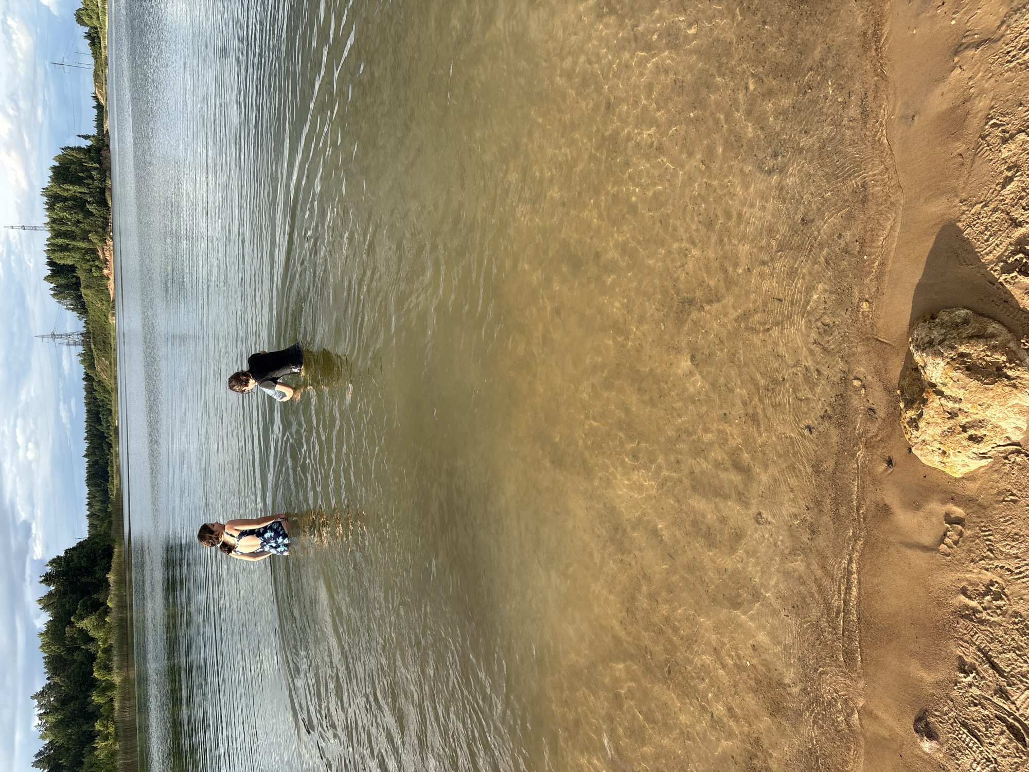 In the city of Zamost'e, Russia, two individuals are wading in a shallow body of water. The person on the left, facing away from the camera and looking towards the right, wears a dark swimsuit with a floral pattern. The person on the right, also facing away and slightly to the left, is clad in a black wet suit or dark-colored long-sleeved garment. The water is clear enough to reveal a sandy bottom. The background features a dense line of trees and a hazy sky with clouds and power lines. The foreground shows a sandy shore with a large, light-colored rock.