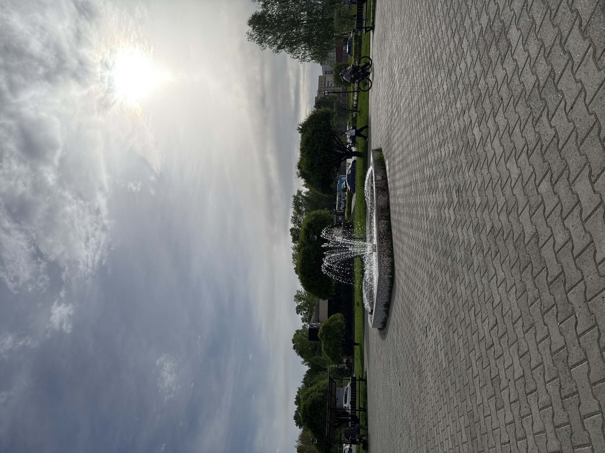 The image depicts a park setting in an unidentified city. A paved walkway, constructed from interlocking pavers, occupies the foreground and extends into the midground. A fountain with multiple jets of water is situated adjacent to the walkway. Beyond the fountain, a strip of grass is lined with trees and parked vehicles. In the background, buildings can be seen. A person is walking near the trees, and a cyclist rides further in the distance. The sky is overcast, with the sun partially obscured by clouds.