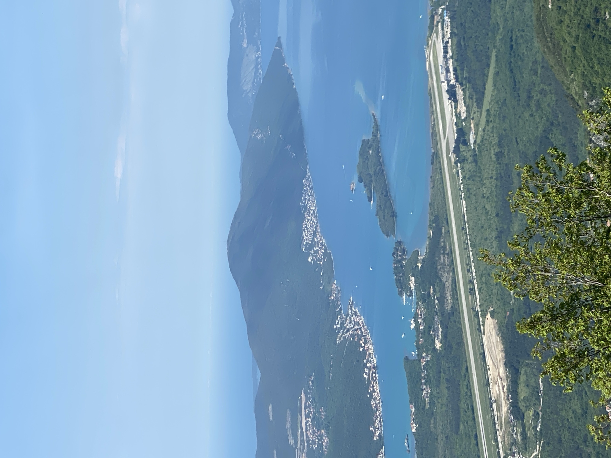 This image presents a stunning, expansive panoramic view from a high vantage point overlooking a mountainous coastal or lakeside region, confirmed by the location context as likely being in Montenegro (Žanjev Do). The scene is dominated by a vast body of clear blue water, perhaps a bay or large inlet, stretching into the distance under a bright, cloudless sky.
Steep, forested mountains and hills flank the water, their slopes covered in dense green vegetation. Along the irregular shorelines and lower mountain slopes, numerous white-roofed buildings form bustling settlements and towns, integrating human activity into the natural landscape. Several small, green islands or peninsulas are visible within the water, some also dotted with buildings. A few small boats are scattered across the blue expanse, indicating leisurely activities or local transport.
A prominent, straight, light-colored strip of infrastructure, resembling either a coastal highway or an airport runway, runs parallel to the water's edge, dissecting the green landscape and connecting some of the coastal communities. In the immediate foreground on the right, a tree branch with fresh green leaves partially frames the vista.
The time of day appears to be bright daytime, likely morning or early afternoon, given the clear illumination and the vibrant blue sky. The weather is sunny and clear, contributing to the tranquil and picturesque atmosphere of the scene. There is no visible text in the image. The overall impression is one of a grand, serene landscape where nature and human settlement coexist.