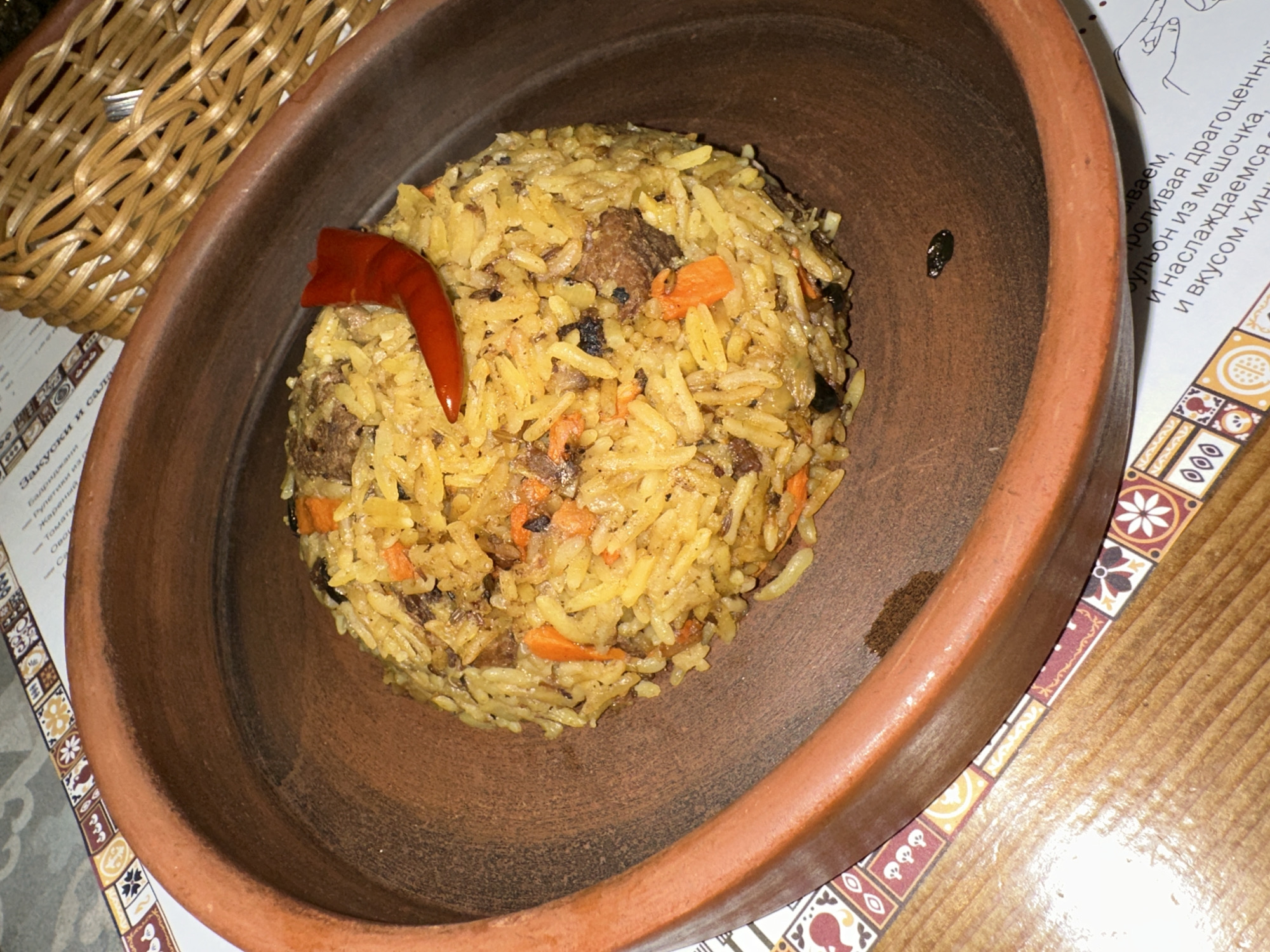 This media file presents a high-angle, close-up view of a dish of pilaf (Plov) served in a rustic, dark brown clay or ceramic bowl. The pilaf, forming a circular mound, consists of yellowish long-grain rice, mixed with chunks of meat (likely lamb or beef), diced orange carrots, and small dark ingredients, possibly raisins or spices. A vibrant red chili pepper is placed on top of the pilaf as a garnish.
To the upper left, a portion of a light brown woven basket is visible. The bowl rests on a patterned paper placemat or menu, which features ornate borders with geometric and stylized floral designs in shades of red, brown, and off-white. The placemat itself is laid on a wooden table surface. A small, dark, oblong object is noticeable on the inner rim of the ceramic bowl, to the right of the pilaf.
The setting appears to be an indoor dining establishment, such as a restaurant or cafe, given the formal presentation and menu. The lighting is somewhat dim, suggesting an evening meal or a cozy ambiance.
Visible Russian text on the left side of the placemat includes menu items such as "Закуски и сала" (Snacks and salads), "Бадриджани" (Eggplant), "Рулетики из" (Rolls of), "Жареный" (Fried), and "Томати" (Tomatoes). On the right, instructional text is partially visible, mentioning "роливая драгоценный" (pouring precious), "бульон из мешочка," (broth from a bag/pouch), "и наслаждаемся" (and we enjoy), and "и вкусом хин" (and the taste of khin, likely referring to Khinkali).
No people are visible in the image. The scene depicts a prepared meal, possibly awaiting consumption. The location context is Saratov, Russia.
