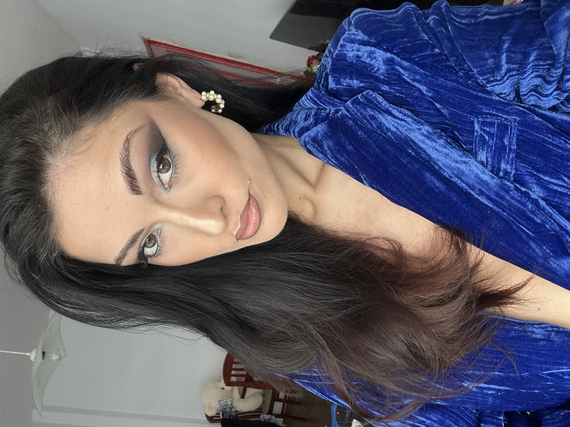 The image is a portrait of a person with long, dark hair, wearing a blue velvet garment. They have fair skin, dark eyebrows, and light-colored eyes with makeup. They are also wearing pearl earrings. The background appears to be an indoor setting with furniture and other objects partially visible. The location and city are unknown.