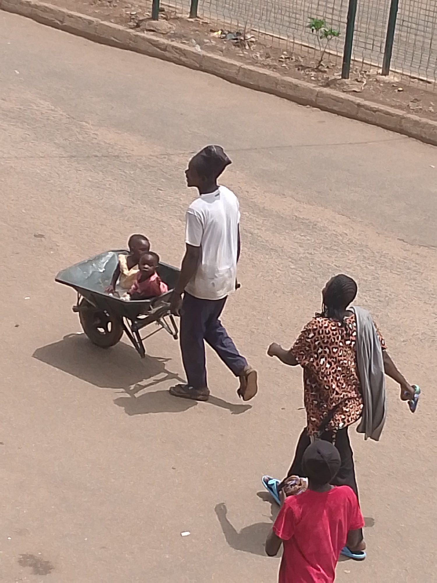 An overhead shot shows four people, one carrying a wheelbarrow containing two children, on a paved road in Bukuru, Nigeria. A person in a white t-shirt and blue trousers pushes the wheelbarrow. The children in the wheelbarrow wear light-colored shirts. To the right of the wheelbarrow is a person in a brown leopard-print shirt, gray shawl, and black pants, holding a pair of blue sandals. A child in a red t-shirt and black pants also wears blue sandals. A metal fence and vegetation are visible on the right side of the photo.