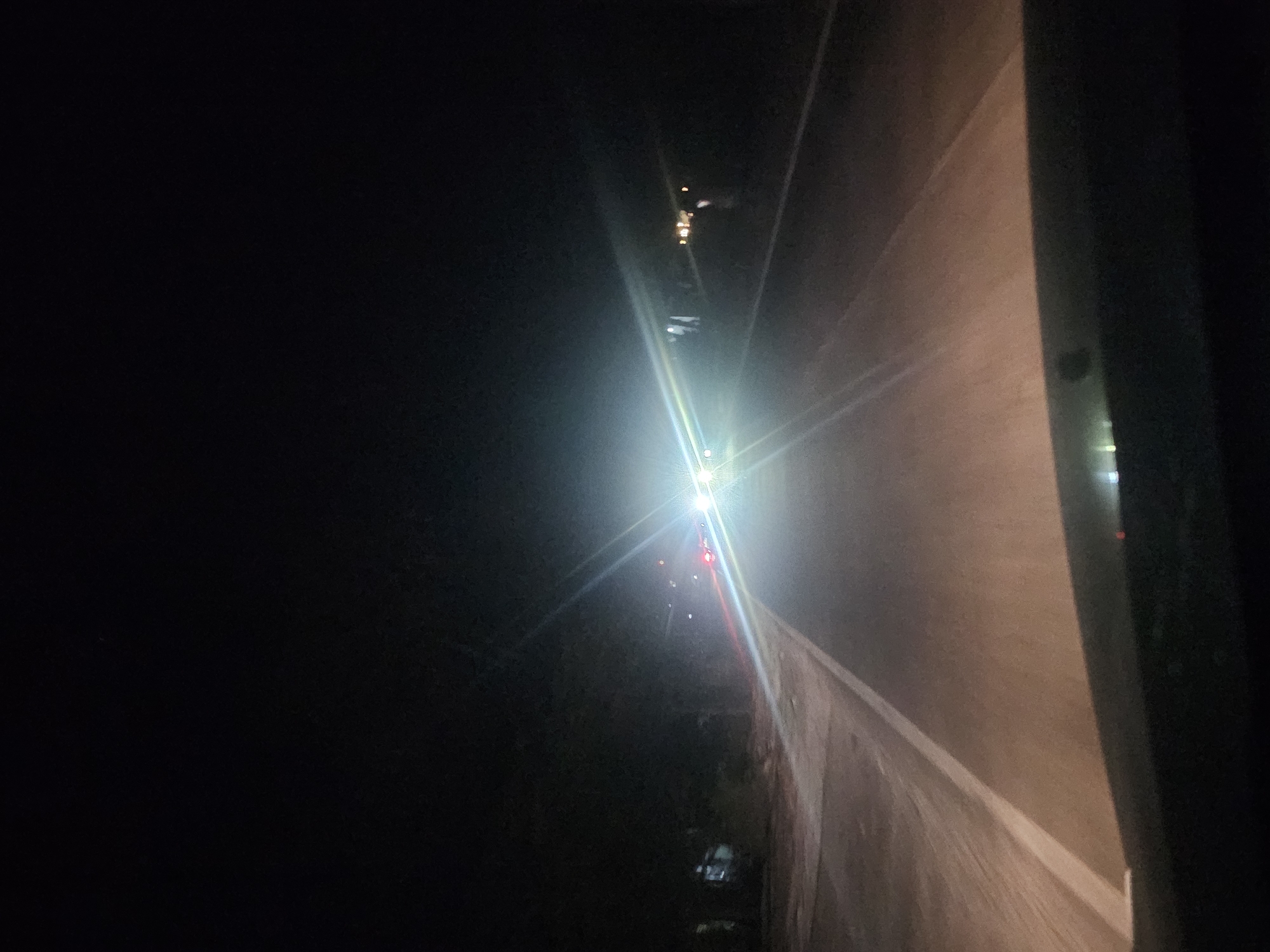 The image depicts a nighttime view from a vehicle driving on a paved road in Sumberwudi, Indonesia. The road has a white line marking the lane. The driver's perspective shows the bright headlights of oncoming vehicles illuminating the scene. Trees and some structures are visible on the sides of the road, shrouded in darkness. The sky is completely black, indicating a very dark night. The foreground of the image is occupied by the dark silhouette of the vehicle's dashboard or hood.