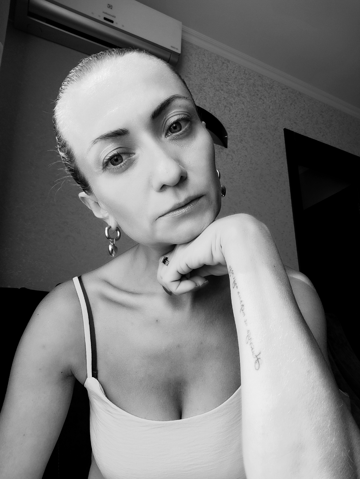 A black and white, medium shot, waist-up selfie taken indoors shows a woman leaning her chin on her hand. Her hair is pulled back tightly, and she is wearing a white tank top with dark straps. She has large hoop earrings with heart-shaped charms. There is a tattoo on her right forearm. She is looking directly at the camera with a neutral expression. In the background, an air conditioning unit is visible on the wall above her. The overall environment appears to be residential.