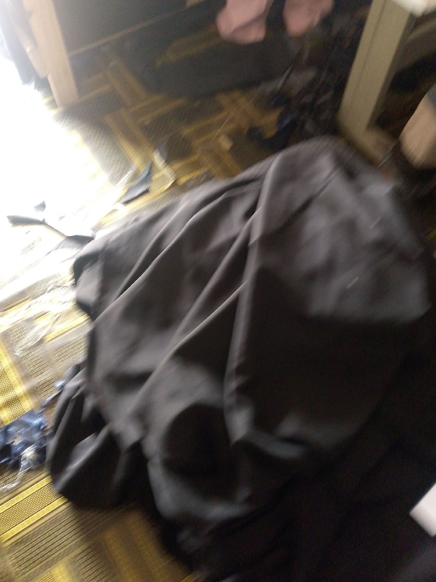 The image is an interior scene, likely a room, with a floor covered in a patterned mat. A large, dark, draped fabric dominates the foreground, appearing to be bunched up and slightly messy. To the left and slightly behind the fabric, a bright light source is visible, casting a strong glare. The background is blurred and indistinct, showing hints of furniture and possibly other fabric items. There are no people visible in the image, and no specific objects or actions are clearly discernible due to the blurriness and focus on the fabric. The location in the city of Jauro Yinu, Nigeria, is not visually indicated in the image.