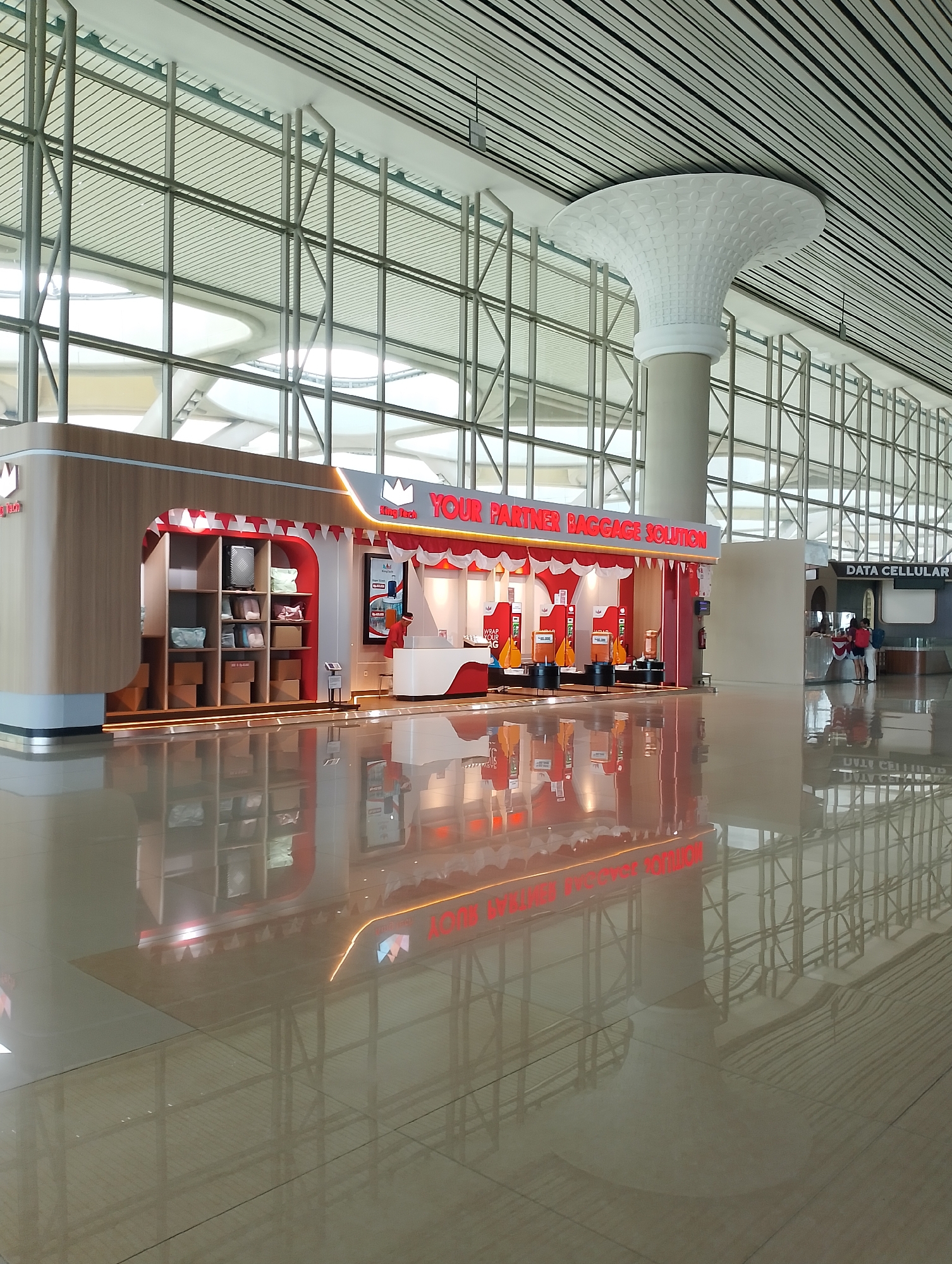 This is a photograph of the interior of an airport terminal in Palihan, Indonesia. The terminal features a modern architectural design with a high ceiling supported by metal beams and a reflective, polished floor.
In the foreground, a well-lit kiosk is prominently displayed, branded with "King Tech" and "YOUR PARTNER BAGGAGE SOLUTION." The kiosk has a wooden exterior and a white and red interior, displaying various suitcases and bags on shelves. There are also automated machines for wrapping luggage.
To the right of the baggage solution kiosk, there is a "DATA CELLULAR" store with customers interacting at the counter.
The floor of the terminal is highly reflective, mirroring the architecture and the kiosks, creating a sense of depth and spaciousness. The overall atmosphere appears clean and functional, typical of a modern airport environment. The lighting is bright and diffused, highlighting the contemporary design elements.