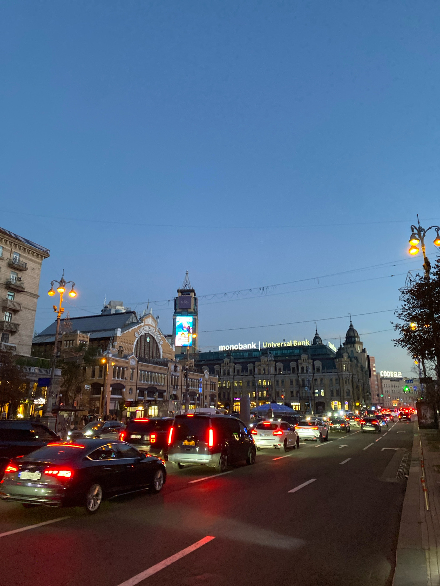 This image shows a street scene in a city at dusk. The sky is a deep blue, and the streetlights are on, casting a warm glow on the traffic. A line of cars is moving down the street, their headlights and taillights illuminating the road. Buildings line the street, some of which are lit up with lights. In the background, a large billboard displays an advertisement. The overall impression is one of a busy city street at the end of the day.