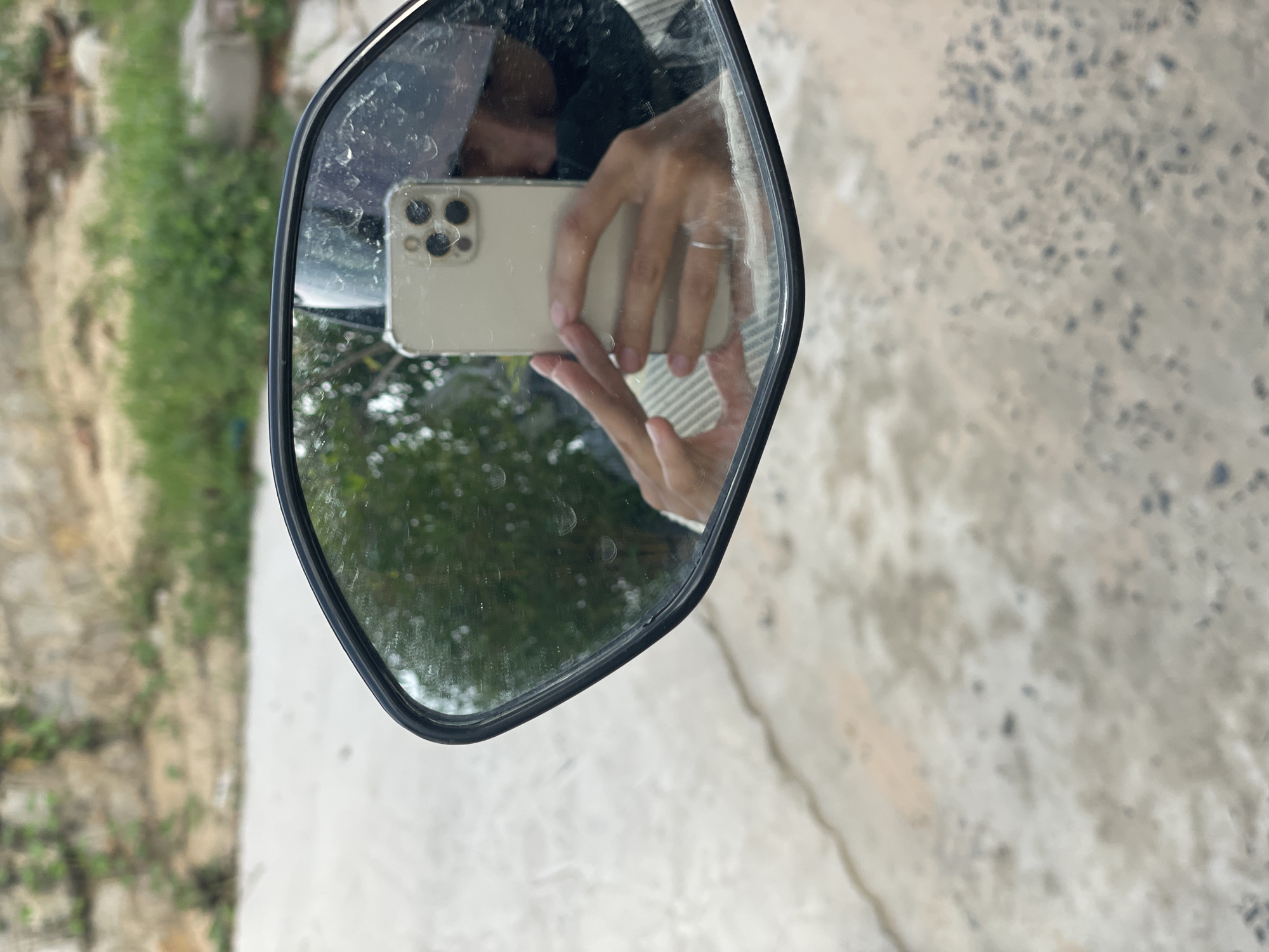 An outdoor, eye-level shot captures a person taking a selfie in a dirty rearview mirror, likely from a vehicle. The mirror, framed in black, reflects the person's hands holding a light-colored smartphone with a visible triple camera system, suggesting an iPhone model, encased in clear plastic. A silver-colored ring is worn on the ring finger of the hand holding the phone. Part of the person's head or face is vaguely visible above the phone.
The mirror's surface is smudged and covered with numerous small specks, possibly dirt or water residue. The reflection within the mirror shows greenery and what appears to be an outdoor path or street, indicating a daytime setting. Outside the mirror frame, to the right, is a textured concrete or paved ground surface with scattered dark spots. To the left and bottom of the mirror, the background is blurred but shows green foliage and a lighter, possibly unpaved, ground area. There is no visible text in the image. The scene is set in Phu Thuy Ward, Vietnam.
