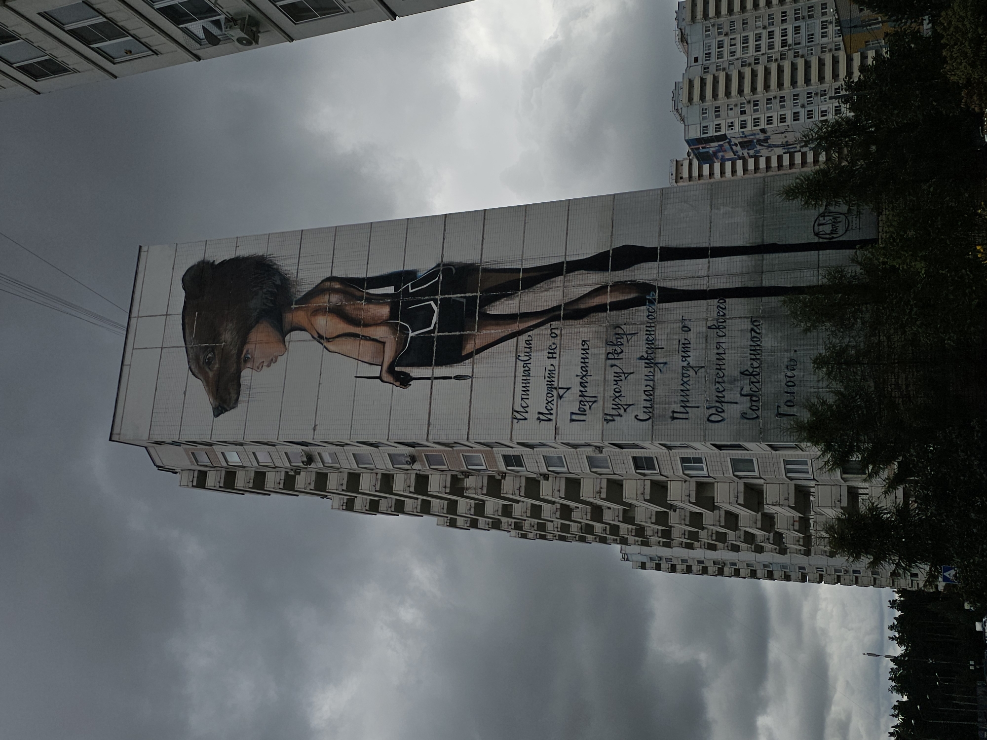 The image shows a tall, multi-story apartment building with a large mural painted on its side in Odintsovo, Russia. The mural depicts a stylized young person wearing a bear's head as a hat and holding a paintbrush. The figure is rendered in shades of brown and black, with a pale complexion. Text in Russian is painted vertically alongside the figure. The building itself is white with numerous balconies, and other apartment buildings are visible in the background. The sky is overcast with dark clouds. Trees and streetlights are present at the base of the building.