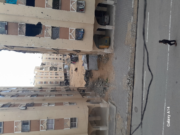 An elevated view captures a damaged urban environment in Khan Yunis, Palestinian Territory. The scene is dominated by multiple multi-story residential buildings with light-beige facades and brown trim. The two closest buildings, flanking a central sandy lot, exhibit extensive damage, including numerous broken and missing windows, some covered by tarpaulins or fabric, and large spall marks or holes on exterior walls. Decorative arched window surrounds are visible. The ground floor of the right building has open arched areas with a yellow tarp-covered object and a light-colored vehicle parked beneath. The central sandy lot is strewn with debris, rubble, and features several small, makeshift shelters with tarpaulin coverings; laundry hangs near one shelter. In the background, additional damaged residential structures are present. In the foreground, an asphalt road with white markings is visible. A single individual in dark clothing walks across the road from right to left. Overhead utility wires traverse the upper foreground. The sky is overcast. A 'Galaxy A14' watermark is present in the lower-left corner of the image.