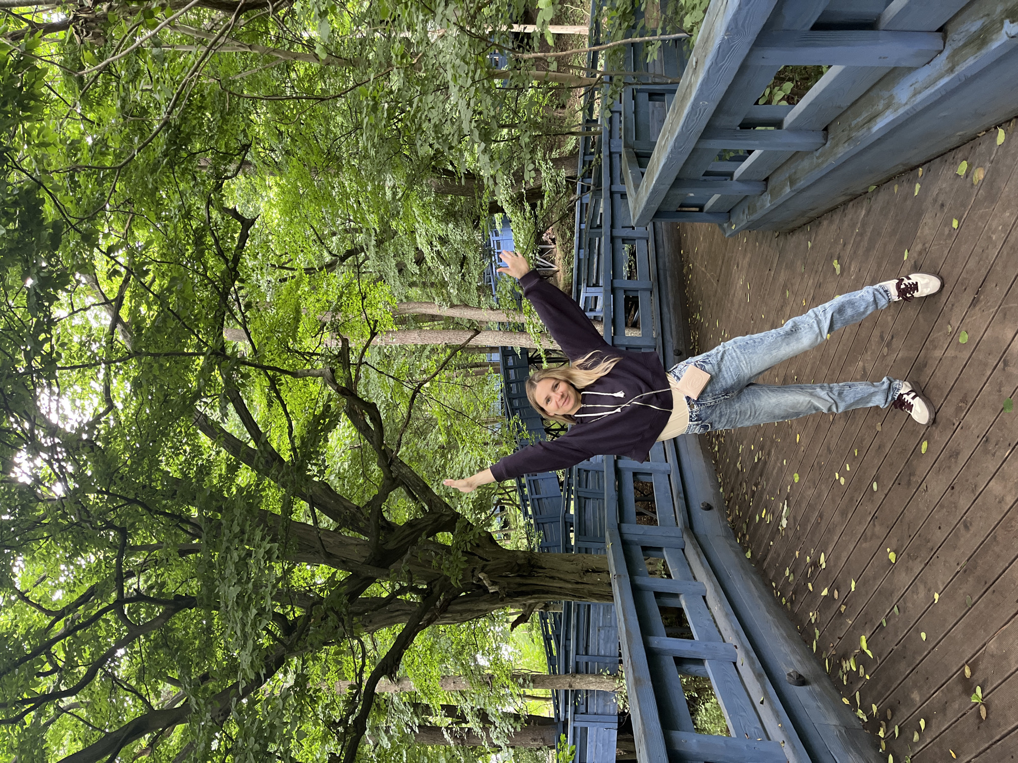 A young woman stands on a wooden boardwalk with blue railings, her arms raised in a gesture of greeting or excitement. She is wearing a dark hooded sweatshirt, a light-colored crop top, light-wash jeans, and sneakers. A small light pink crossbody bag is slung over her shoulder. The boardwalk curves through a lush green forest, with a large, gnarled tree prominently featured on the left. Sunlight filters through the dense canopy of leaves, creating dappled patterns on the boardwalk. Scattered green and yellow leaves lie on the wooden planks. The background shows more of the elevated blue boardwalk winding through the trees. The scene suggests an outdoor recreational area in a wooded environment, potentially a park or nature trail. No specific landmarks of Pionersky, Russia are identifiable in the image.