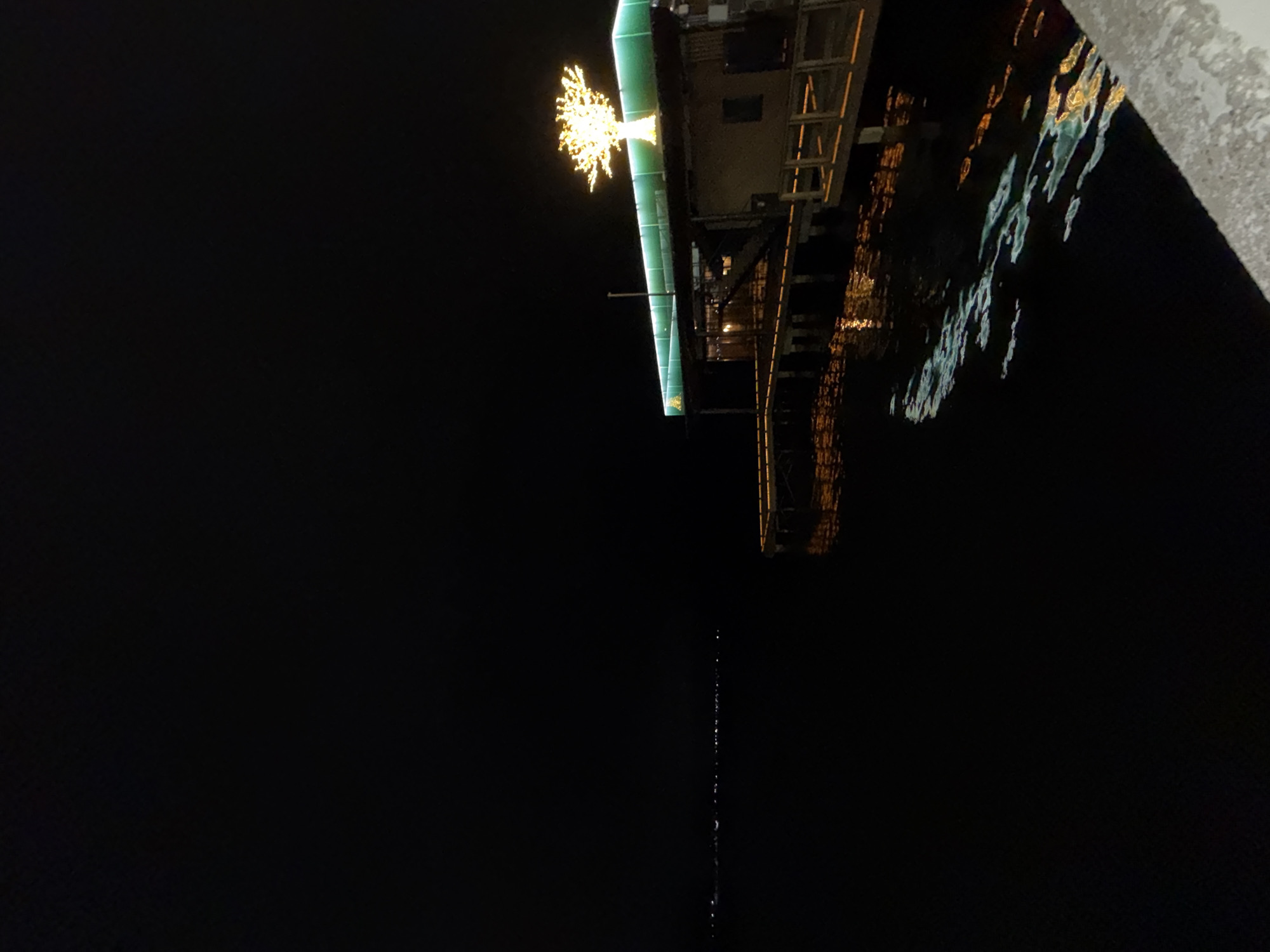 The image captures a nighttime scene in Votkinsk, Russia, showing a brightly lit structure extending over dark water. The perspective is looking down and across, with the structure positioned in the upper right quadrant, angled slightly.
The most prominent feature is a radiant, golden-yellow light fixture shaped like an abstract star or a small, illuminated tree, fixed onto the side of a building or pier. This structure appears to have a green-colored edge or beam along its length, and other warm, smaller lights are visible on its underside or within its framework.
The dark water below reflects these lights, creating shimmering patterns of gold, yellow, and subtle blue-green. The reflections are somewhat distorted, indicating gentle movement in the water. A distinct, very thin vertical line of small, bright dots or reflections extends far down into the black expanse of the water from near the structure, suggesting a distant or narrow light source reflecting on the surface.
In the upper right corner of the foreground, a textured white surface, possibly snow, ice, or a concrete edge, frames part of the view. The overall scene is devoid of people or discernible activities, focusing on the illuminated architecture and its reflections. The time of day is clearly night, characterized by deep darkness. There is no visible text in the image.