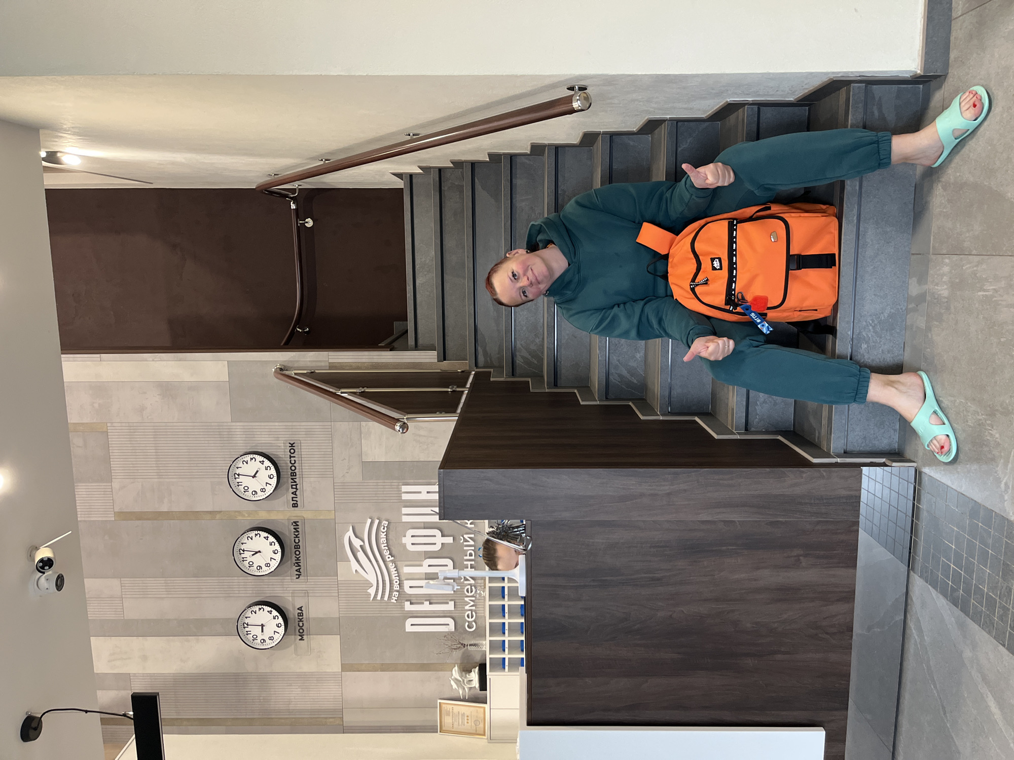 A woman sits on the stairs in front of the "Dolphin" family complex in Chaikovsky, Russia. She is wearing a green sweatsuit and light green sandals and has an orange backpack behind her. She is giving a thumbs-up sign with both hands. Behind her is a wall with three clocks showing the time in Moscow, Chaikovsky, and Vladivostok. There is also a logo for "Dolphin" on the wall. A security camera is visible on the wall to the left. A staircase with a wooden handrail is visible.