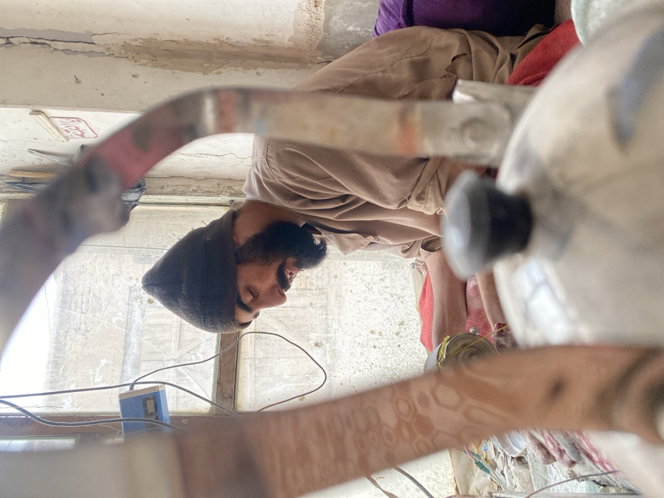 An indoor scene in Kandahar, Afghanistan, depicts a single male individual engaged in a focused task. The man, seen in profile facing right, has a dark beard and wears a dark knitted cap and a light brown, long-sleeved garment. He is seated on the floor or a low surface, leaning forward, with his hands actively working on an object in his lap, which includes visible yellow material, possibly thread or wire.
In the immediate foreground, out of focus, a silver-colored kettle with a black knob on its lid partially obstructs the view. Curved, out-of-focus elements, likely parts of the kettle or another foreground object, frame the central figure: one on the left shows a hexagonal pattern, and another on the right has red and grey markings.
Behind the man, a window with wooden framing offers a blurred view of an exterior wall or structure. Electrical wires are strung across the window area, connecting to a light blue electronic device mounted nearby. The interior walls show signs of wear, including cracks and peeling paint. A small white rectangular sign with red Arabic script ("يا ألله" - Ya Allah) is affixed to the wall above the man's head. To his right, a red fabric is visible, and to his left, a purple cushion or fabric. The environment appears utilitarian, illuminated by natural light entering through the window.