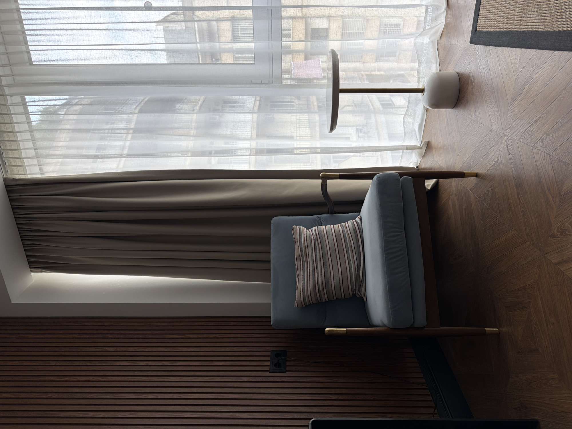 The image shows an interior scene, likely a living room or lounge area, within a city. The style suggests a modern and minimalist aesthetic.
In the foreground, a dark brown wooden floor with a parquet pattern is visible. To the left, a textured dark brown wooden panel covers the wall, featuring vertical slats. An electrical outlet is mounted on this panel.
Dominating the center-left is a light blue armchair with wooden legs and arms, adorned with a striped cushion. To the right of the armchair, a small, round-topped side table with a white base and a slender metal stand stands near a large window.
The window is covered by two layers of curtains: a thicker, taupe-colored drape on the left and sheer white curtains on the right. Through the sheer curtains, the facade of a multi-story building with balconies and windows is visible, suggesting an urban environment.
The overall lighting is soft, indicating either natural light filtering through the curtains or interior lighting. There are no people visible in the image.
While the image depicts an interior setting with a view of a city building, there is no specific visual information within the image that allows for the definitive identification of the city as Tbilisi, Georgia. The architectural style of the building outside is common to many European cities, and the interior design elements are not uniquely Georgian.
