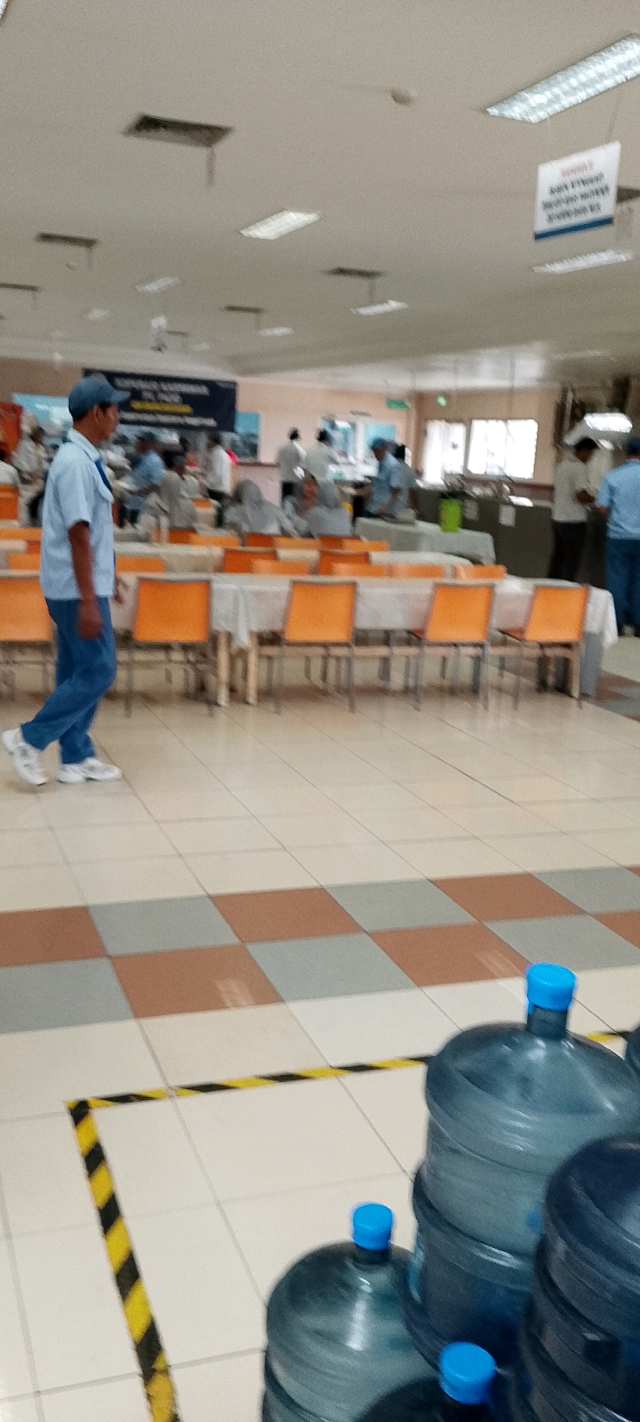 The image depicts an indoor scene, likely a cafeteria or dining hall within a facility in Depok, Indonesia. In the foreground, a man wearing a light blue shirt, dark blue pants, and a blue cap is walking from left to right. He is wearing white sneakers. The floor is tiled in a checkered pattern of white, beige, and brown squares. Yellow and black hazard tape is visible on the floor in the lower left quadrant.
In the midground, several long tables are set with white tablecloths. Orange chairs are arranged around the tables. In the background, various people are seen standing and seated at the tables, some appearing to be eating or in conversation. Some individuals are wearing headscarves. There are also several large blue water cooler bottles stacked in the lower right corner of the image.
The ceiling is visible at the top of the image and features fluorescent lighting fixtures and what appear to be air conditioning vents. A dark banner with white text is hung on the wall in the background. The overall impression is that of a communal eating area in a workplace or institution.