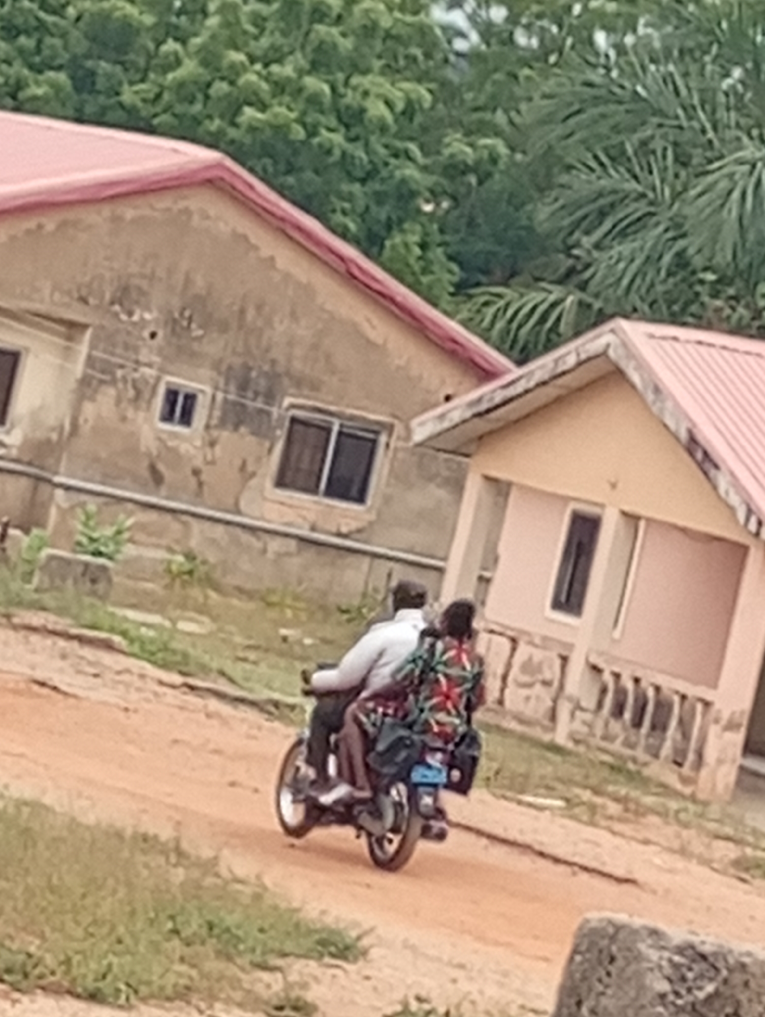 A slightly blurred image captures two people riding a motorcycle on a dirt road. The man driving is wearing a white shirt and dark pants, while the passenger behind him is clad in a patterned floral top. Behind them, there are two single-story buildings with red roofs. The left building appears older, with a weathered exterior, while the one on the right looks more recently painted, featuring pillars and a window. The surroundings are lush with green trees and foliage. The location is presumed to be in a non-urban setting.