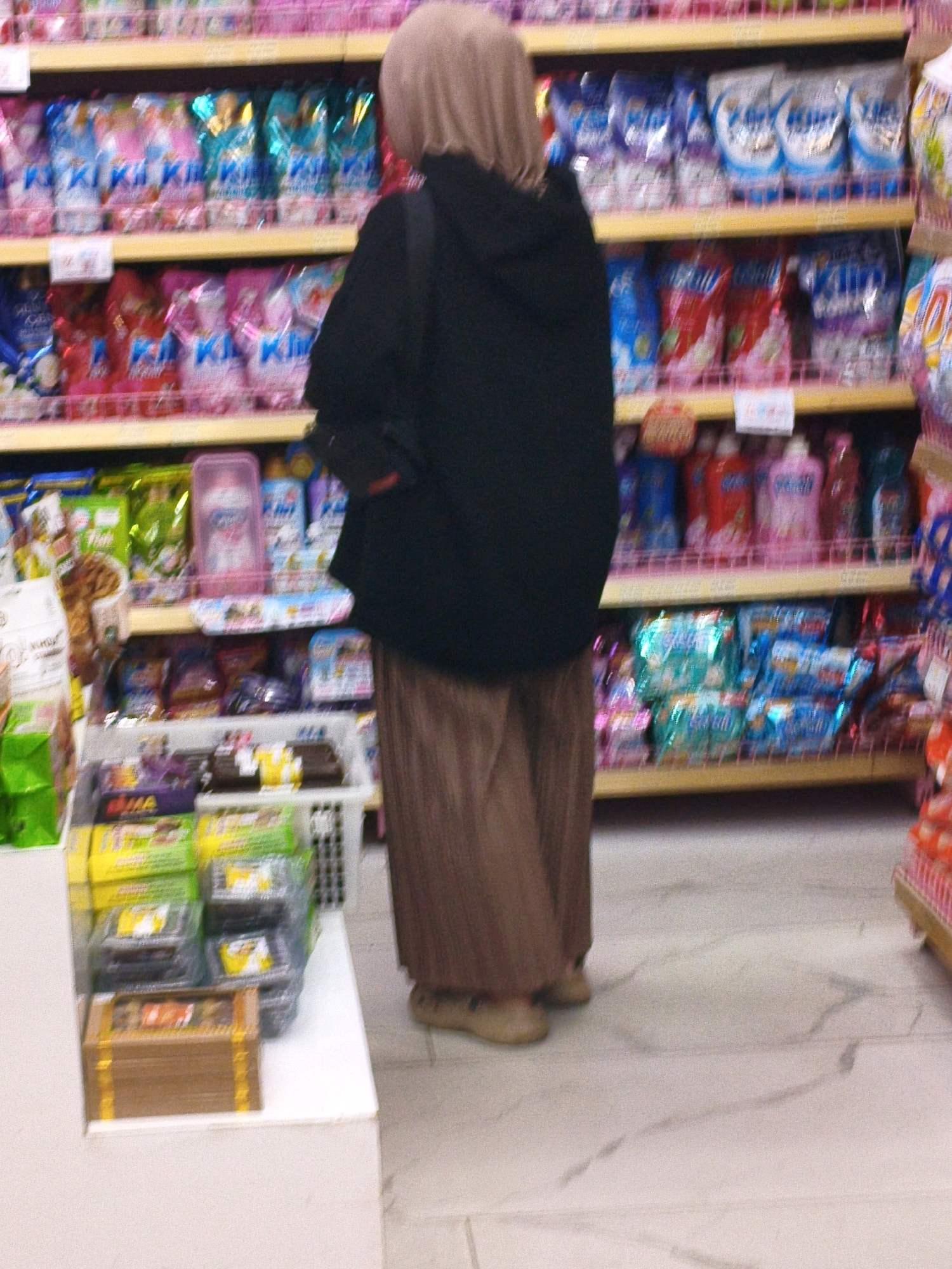 In an indoor environment, a person wearing a beige hijab, a black jacket, and brown pants stands in an aisle of a store, facing away from the camera. The person is positioned in front of shelves stocked with various retail products, primarily cleaning supplies and toiletries. The shelves are brightly lit and organized with numerous colorful packages. In the foreground, a white display unit holds several boxes and packaged items, including what appear to be chocolates or candy bars. The floor is a light-colored, reflective surface, possibly marble or tile. The scene appears to be in a supermarket or department store.