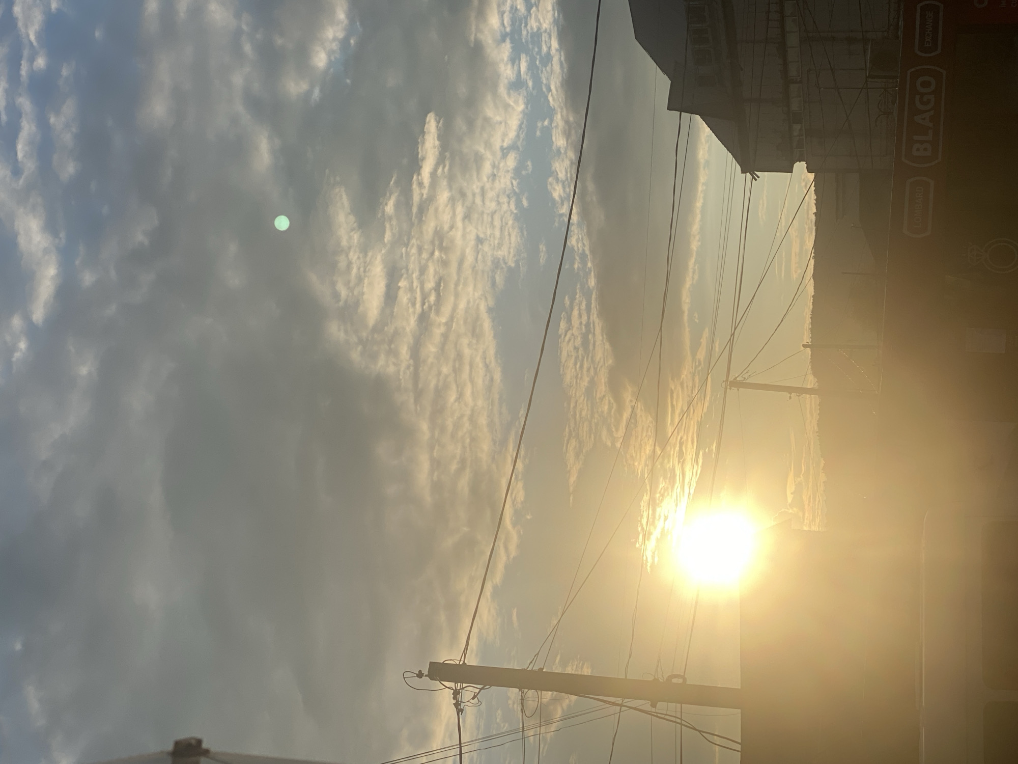 The image captures a bright sunset over the city of Otaci, Moldova. The sun, a dominant feature, is low in the sky and intensely luminous, casting a warm, golden glow across the scene. Its light diffuses through a cloudy sky, where scattered, wispy clouds are illuminated by the setting sun, creating a textured and dynamic atmosphere. A noticeable green orb, possibly a lens flare or atmospheric phenomenon, is visible in the upper right quadrant of the sky.
In the foreground, a utility pole with numerous wires is prominent on the left side, silhouetted against the bright sky. The wires crisscross the frame, adding a sense of urban infrastructure. To the right, the dark silhouette of a building emerges, its upper floors partially obscured by the glare of the sun. Below this building, a red sign with white lettering is visible, displaying words like "LOMBARD" and "BLAGO EXCHANGE," indicating a commercial area within the city. The lower portion of the image is dominated by a bright, hazy light, suggesting the presence of a vehicle or structure that is reflecting or bathed in the sunset's radiance. In the background, a hilly landscape is dimly visible, fading into the warm haze. The overall impression is one of a vibrant, late-day urban environment under a dramatic sky.