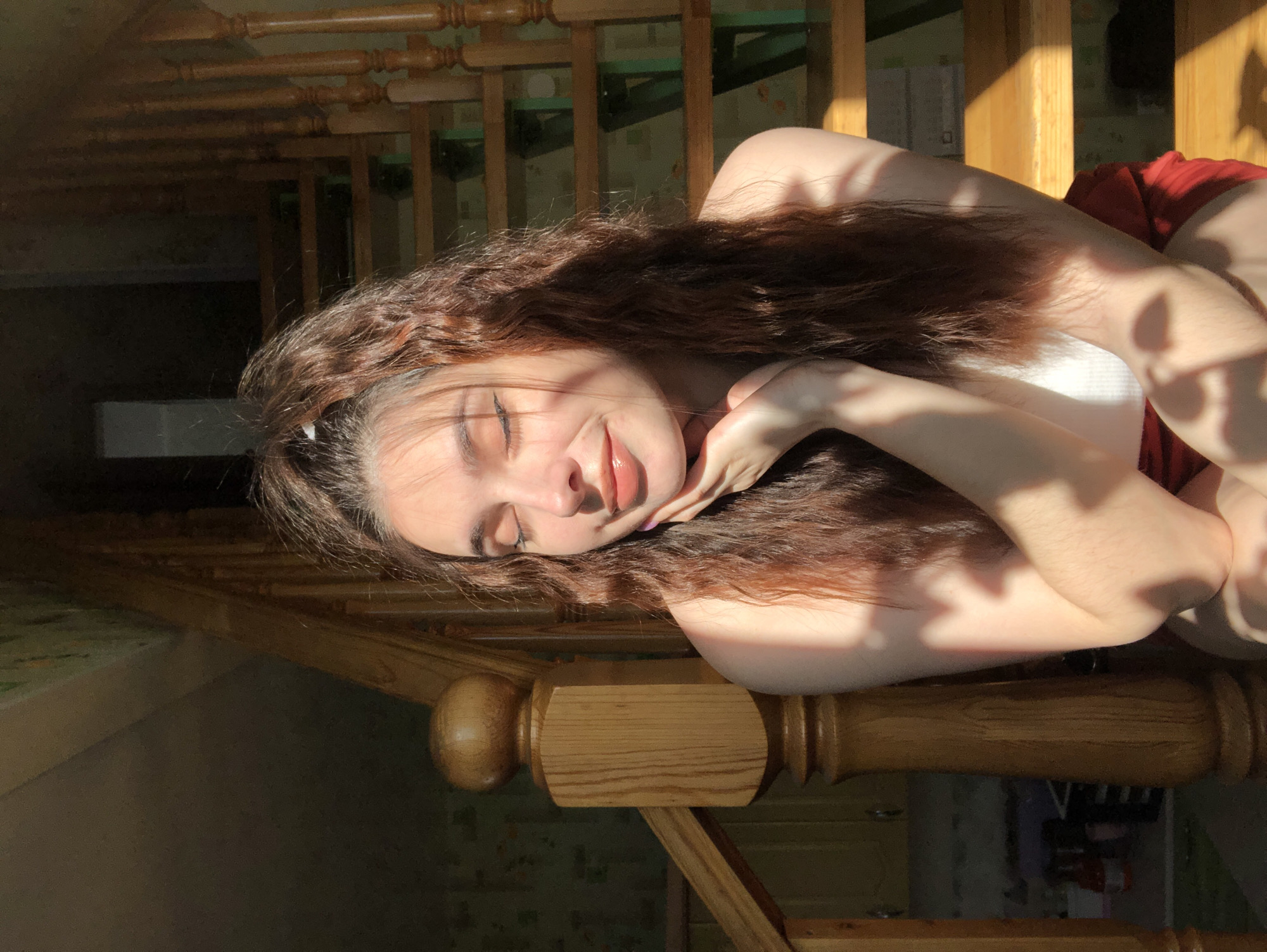 A woman is lying on a wooden staircase, likely indoors. She has long, brown, wavy hair and is wearing a white top and a red garment. She is resting her head on her hand. The lighting suggests it is daytime with sunlight streaming in. The background shows other parts of the staircase and what appears to be a window with shadows cast by foliage. There's nothing to indicate the location as the city of Noviy, Russia.