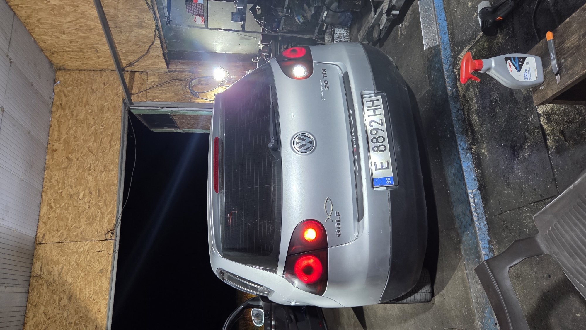 The image shows the rear of a silver Volkswagen Golf parked indoors, possibly in a garage or workshop. The car's brake lights are illuminated. A license plate with the characters "E 8892 HH" is visible, along with a blue and white European Union identifier. The car also has a fish emblem and "GOLF" lettering on the trunk. The background includes wooden paneling, exposed wiring, a fluorescent light fixture, and shelves stocked with various items. In the foreground, a red spray bottle and a utility knife rest on a wooden stool, and a dark plastic object is positioned on the floor.