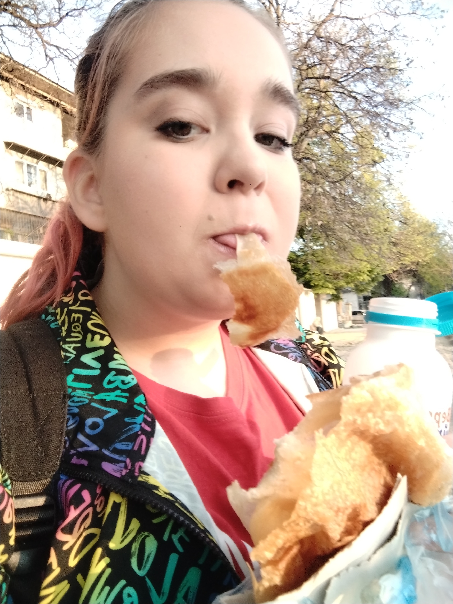 A woman is eating a pastry outdoors, likely in Plovdiv, Bulgaria. She is looking at the camera, holding a large pastry in her hand while eating a bite of it. The pastry appears to be in a white paper bag. She is wearing a red t-shirt and a jacket with colorful letter designs. A brown backpack strap is visible on her shoulder. A white plastic bottle with a blue lid is partially visible. In the background, there is a building and trees. The lighting indicates it is daytime.