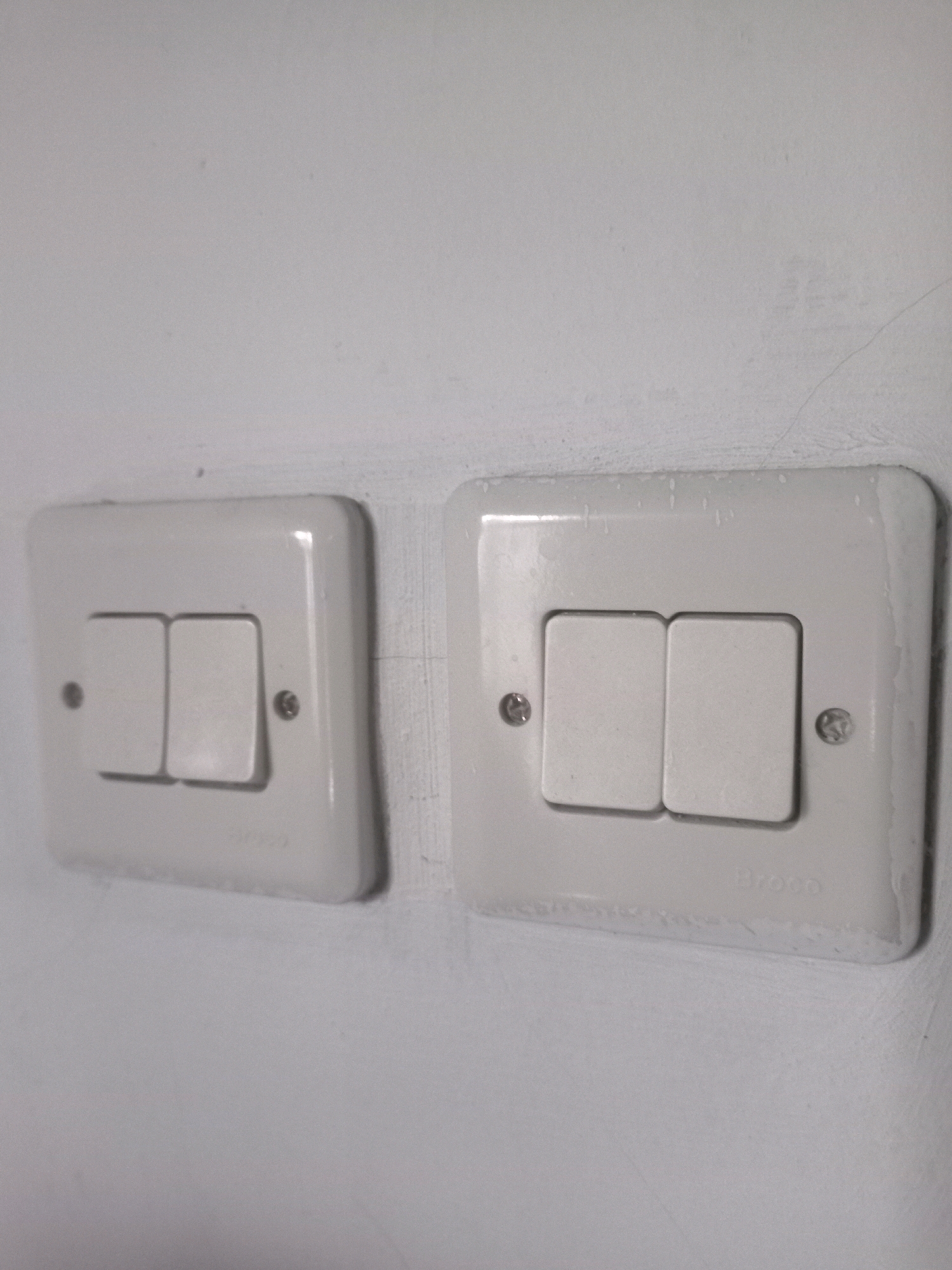 This is a close-up, eye-level shot of two white, double-gang light switches mounted on a textured, off-white wall. The switches are rectangular with rounded corners, and each has two identical rectangular buttons positioned side-by-side. Both switches appear to be in the "off" position, with the buttons facing upwards. The left switch is slightly to the left and lower in the frame than the right switch. The wall shows some minor imperfections, including small dots and faint lines, suggesting it may be plastered or painted. The lighting is diffuse, casting subtle shadows. The brand name "Broco" is visible embossed on the bottom edge of the right switch plate. The image is undated and the specific location in Karangtalun, Indonesia is not visually discernible from the image itself. There are no people or actions depicted.
