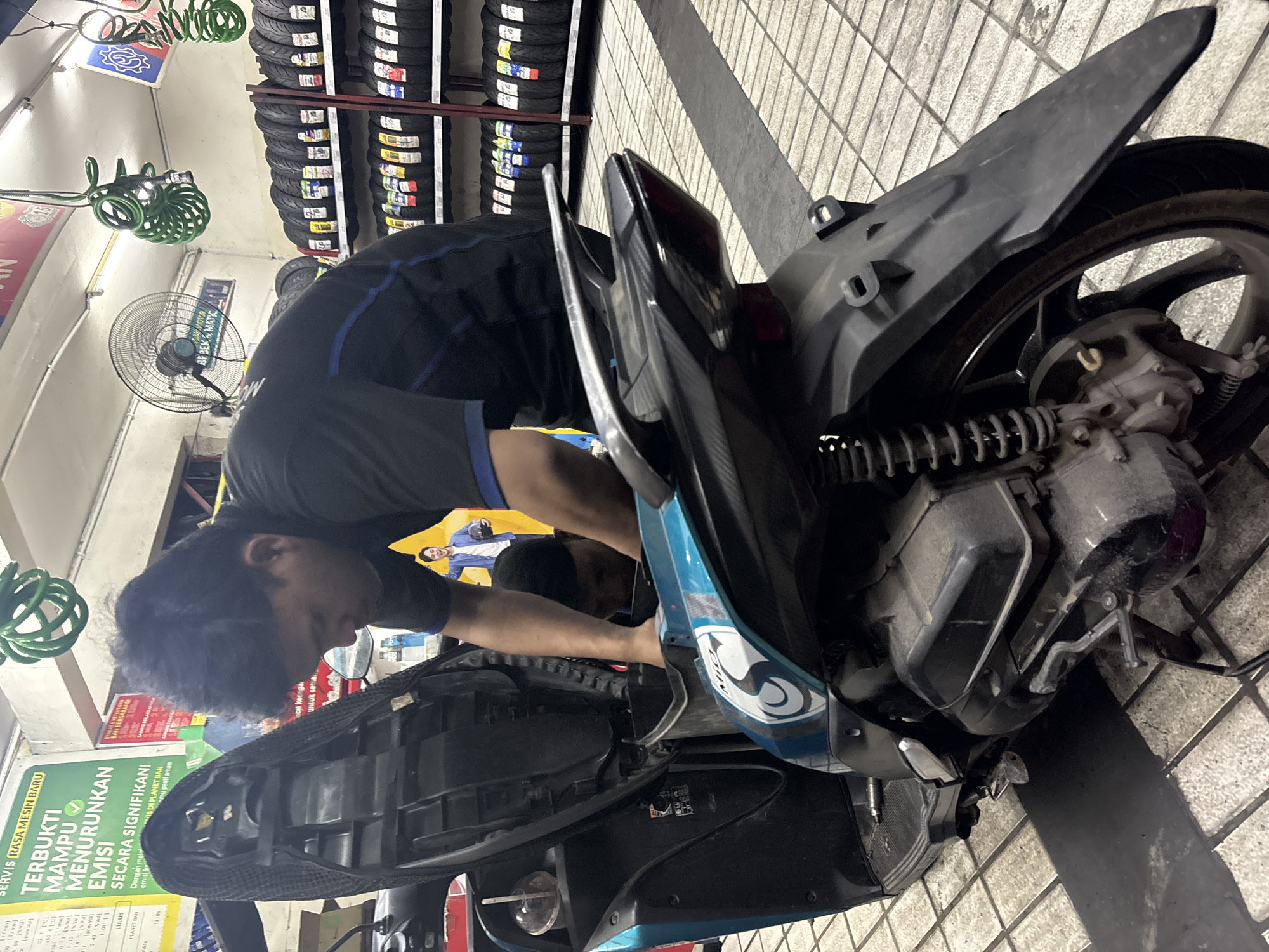 A male mechanic is servicing a teal and black scooter in what appears to be a tire shop in North Jakarta, Indonesia. The mechanic, wearing a black t-shirt with blue accents, is bending over the scooter's open seat compartment, with his head and hands obscured as he works on the engine. The scooter's seat is lifted, revealing the internal components. Shelves stacked with various tires are visible in the background, along with a ceiling fan and coiled green hoses. The floor is tiled with a grid pattern. A large poster featuring a smiling man in a blue shirt holding a helmet is also present. The overall lighting suggests an indoor environment, possibly a garage or a commercial establishment.