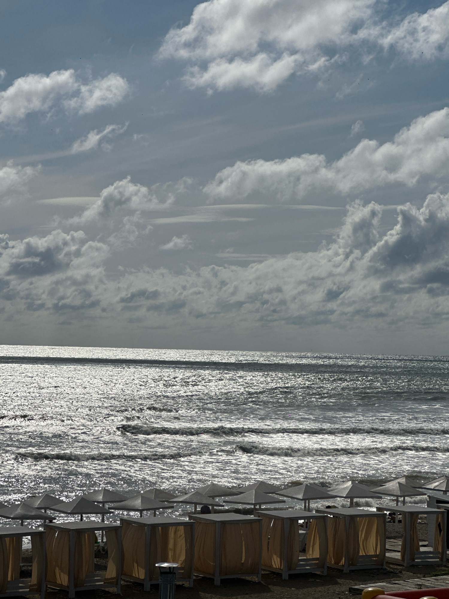 The image shows a beach scene in Lermontovo, Russia, under a cloudy sky. In the foreground, a row of beach cabanas with light-colored curtains are visible. Beyond the cabanas, the sea stretches out, with sunlight glinting off the choppy water. The sky is filled with a mix of white and grey clouds. There are no people clearly visible in the image. The overall atmosphere is serene and somewhat dramatic due to the contrast between the bright sea and the overcast sky.