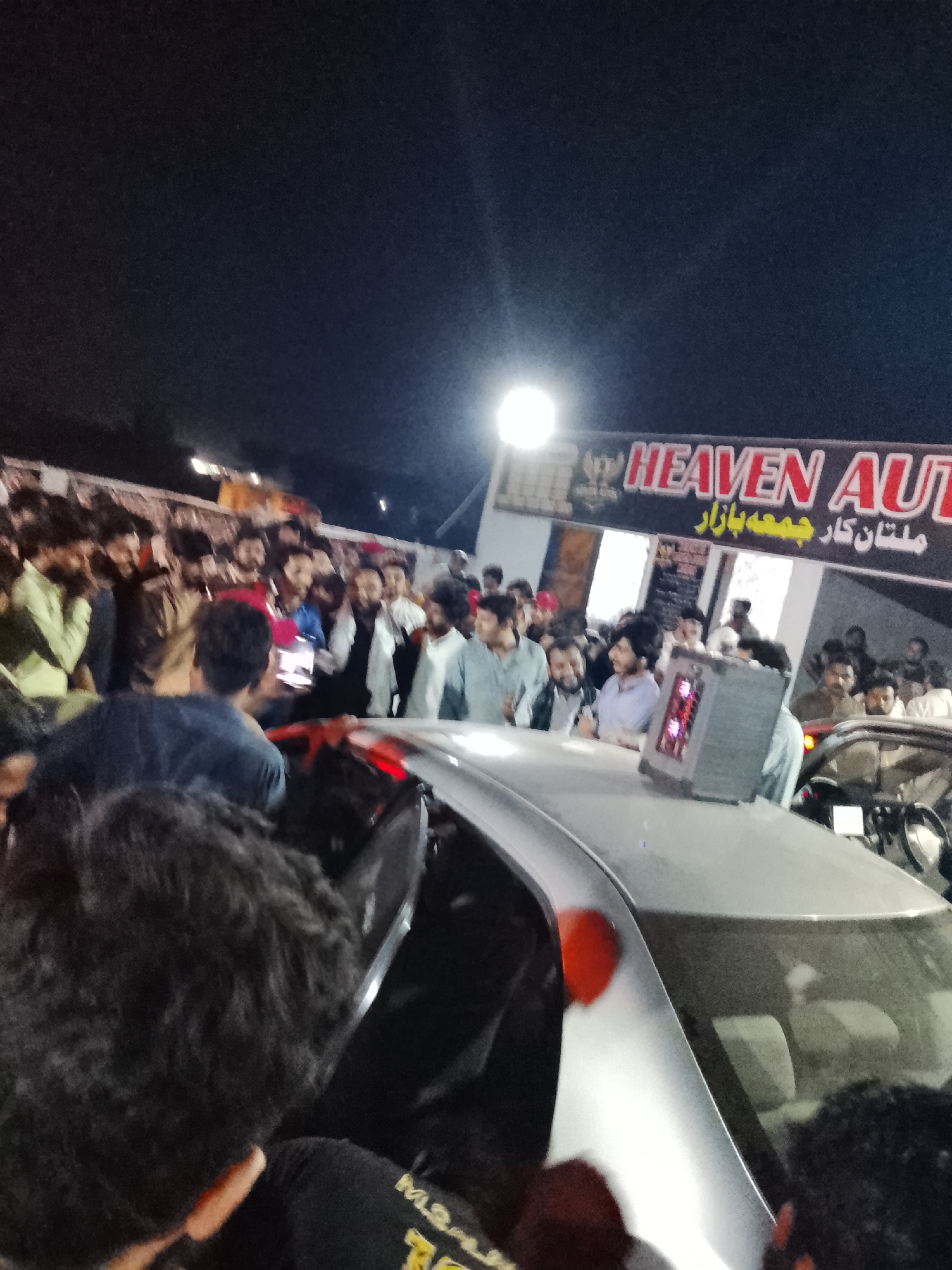 The image captures a nighttime scene at "HEAVEN AUTO ملتانی کار جمعہ بازار", likely an auto bazaar, with a large crowd of people gathered around a silver car. A gray computer tower-like object with red accents sits on the car's roof. Individuals within the crowd are looking towards the car, and some are holding phones, possibly recording or taking pictures. The environment suggests an outdoor location at night, illuminated by artificial light. The location is "ملتانی کار جمعہ بازار". The specific city and region are unknown.