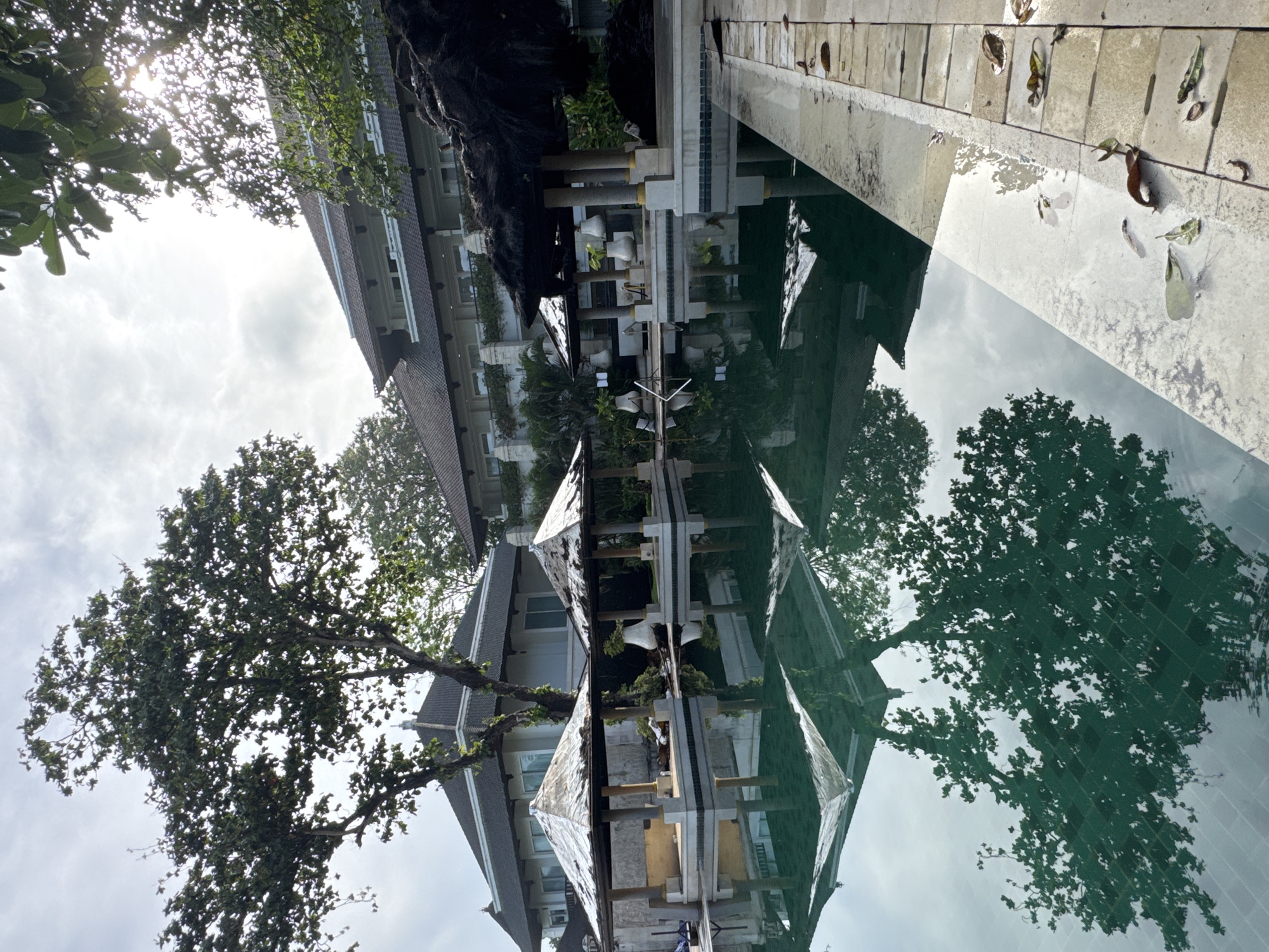 The image, when viewed in its correct upright orientation, captures a serene outdoor scene, likely a resort or hotel grounds in Kuta Selatan, Indonesia. A multi-story building with balconies and dark, possibly tiled roofs occupies the left side of the frame, partially obscured by lush green trees. Sunlight streams through the foliage in the upper left, indicating a bright day, possibly morning or late afternoon, under a cloudy sky.
A large, calm body of water, likely a swimming pool or decorative pond, dominates the right side and foreground. This water perfectly reflects the building, trees, and the bright, overcast sky, creating a strong symmetrical effect. Several decorative pillars or light fixtures, featuring white and gold elements, are positioned along the edge of the water, contributing to the reflections. A light-colored paved area or deck runs alongside the water, scattered with several fallen, dried leaves, some of which are also floating on the water's surface. The scene is devoid of people or specific activity, conveying a sense of tranquility and natural beauty. No visible text is present in the image.