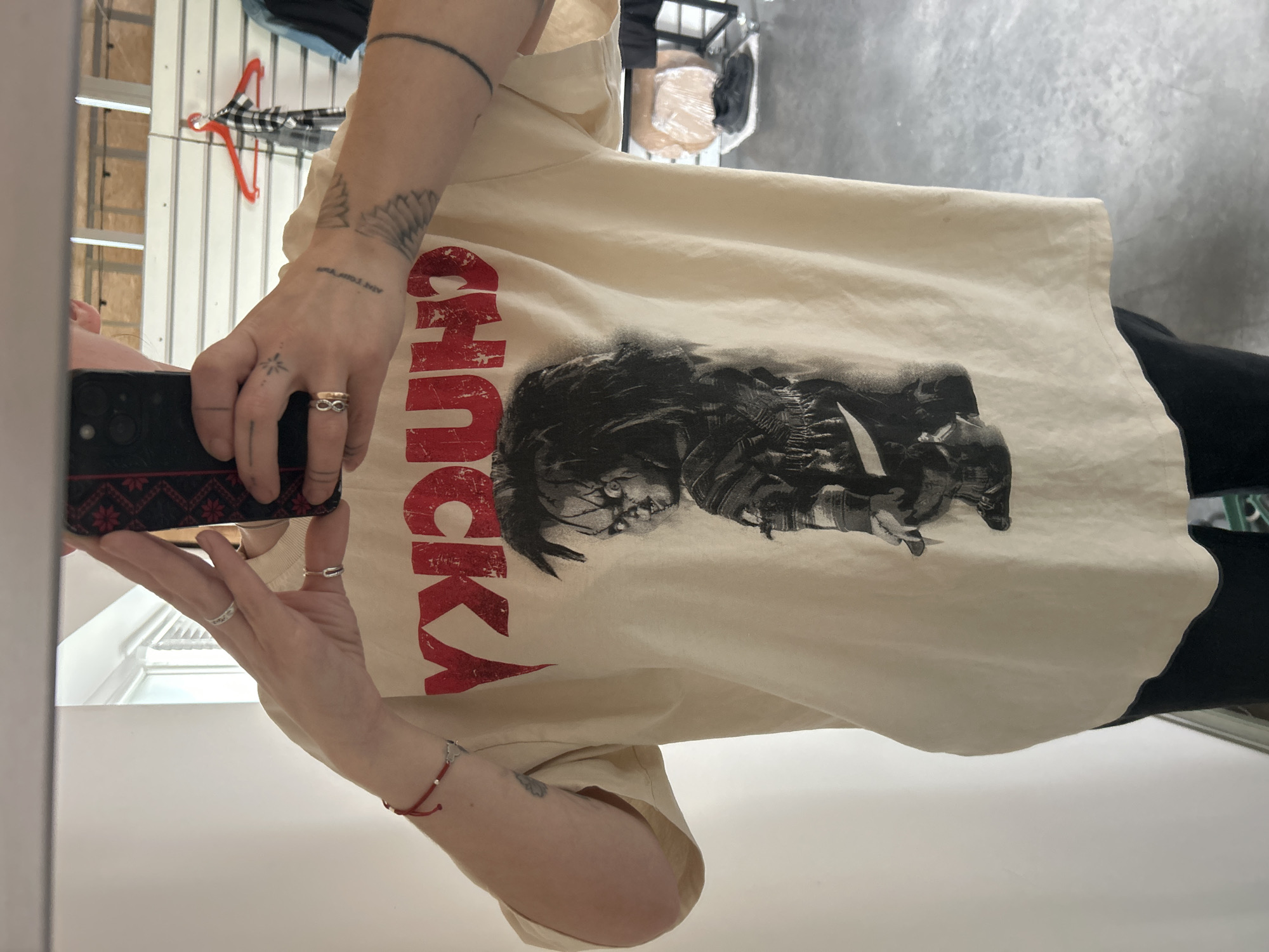 The image shows a person wearing a light-colored t-shirt with a graphic of the character Chucky and the word "CHUCKY" printed on it. The person is holding a smartphone in front of their face, suggesting they are taking a selfie in a mirror. Several rings are visible on their fingers, and a red string bracelet with a small charm is on their left wrist. Tattoos are visible on their right arm and hand. The background appears to be indoors, with shelves and clothing racks visible, indicating a retail or storage environment. The scene does not provide any specific information to determine the location as Ternopil, Ukraine.