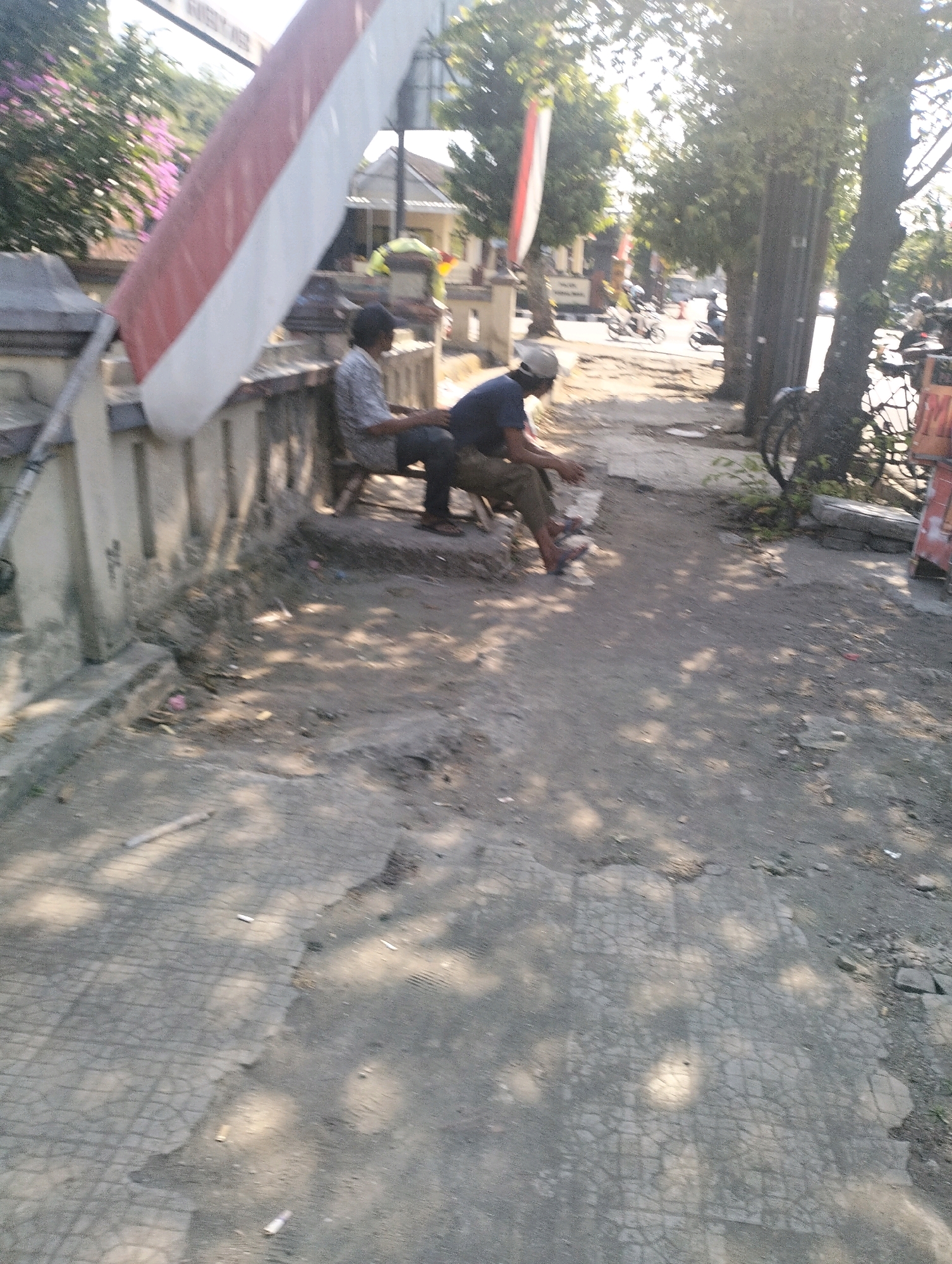 Two men are sitting on a low wall in front of a building on a street in Purbalingga, Indonesia. The man on the left is wearing a light-colored shirt and dark pants and is facing to the right. The man on the right is wearing a blue shirt and dark pants and is facing forward. A large Indonesian flag is propped up behind the men, and a bicycle is leaning against a tree to the right. The ground is paved with concrete, and there are some loose stones and debris scattered around. The sun is shining, casting shadows on the ground.