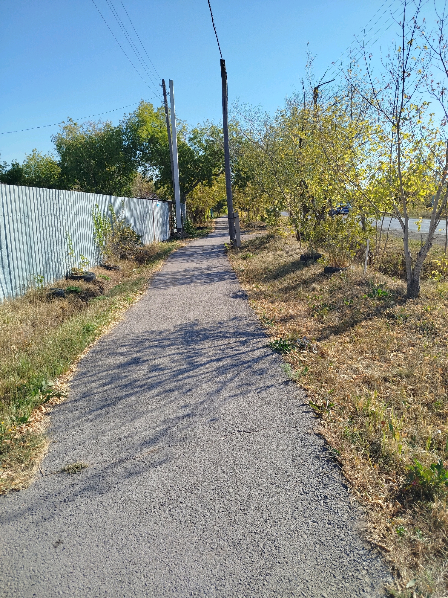 The image shows a paved pedestrian path running alongside a corrugated metal fence on the left and a dry, grassy embankment with trees on the right. The path is shaded by the dappled sunlight filtering through the leaves of the trees, casting linear shadows across its surface. Power lines are visible overhead, supported by wooden poles. In the background, trees with yellowing leaves suggest an autumn season. A dark-colored car is partially visible on a road beyond the embankment on the right side of the image. The scene is set in an outdoor environment with a clear blue sky.