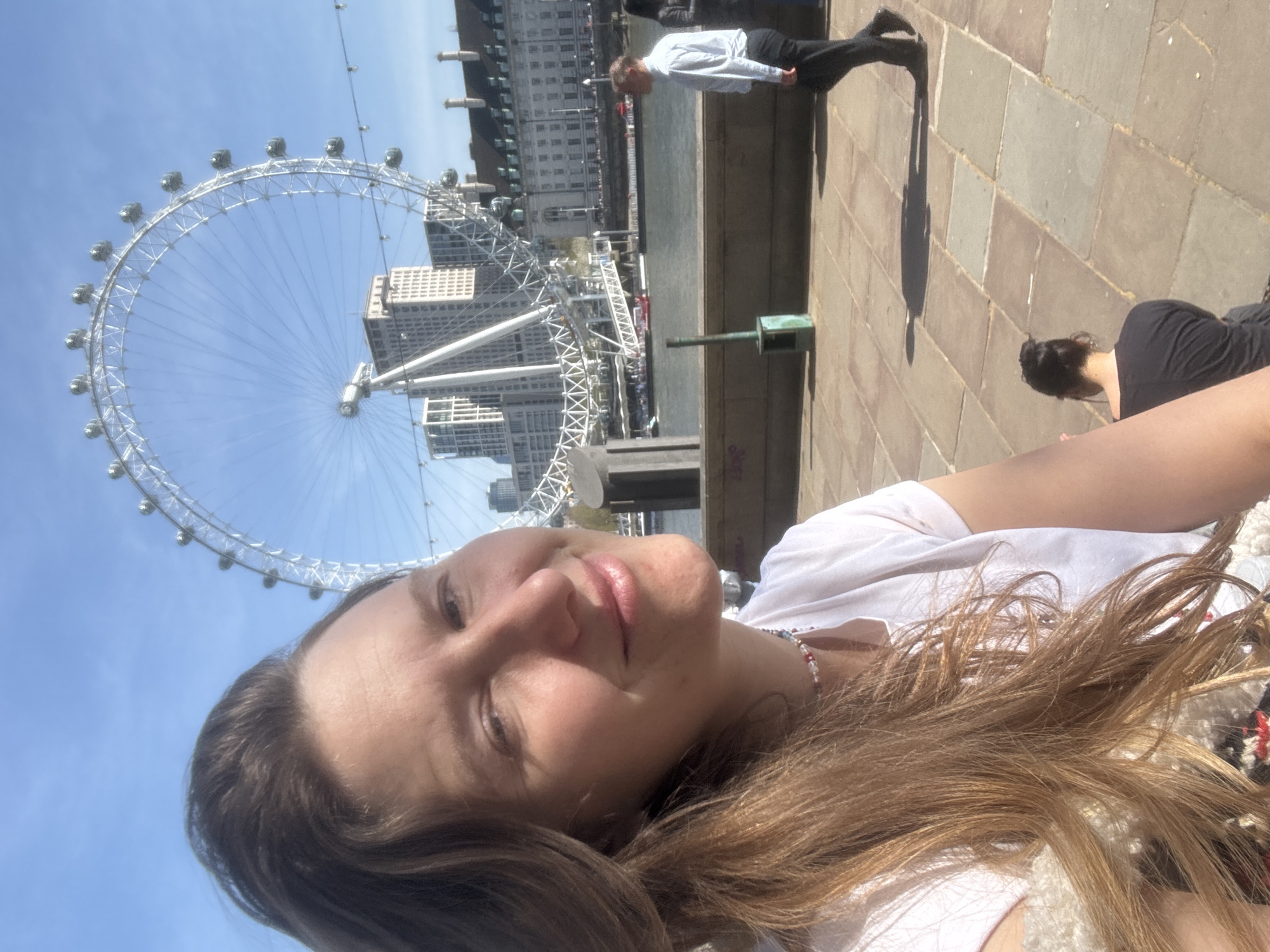 The image shows a selfie of a person with long brown hair. The person is wearing a white shirt and a colorful beaded necklace. In the background, the London Eye is visible, along with the buildings on the opposite bank of the River Thames. A man in a white shirt and black pants is walking on the bank, and another person is kneeling down nearby. The ground is paved with rectangular stones. The sky is blue and mostly clear.