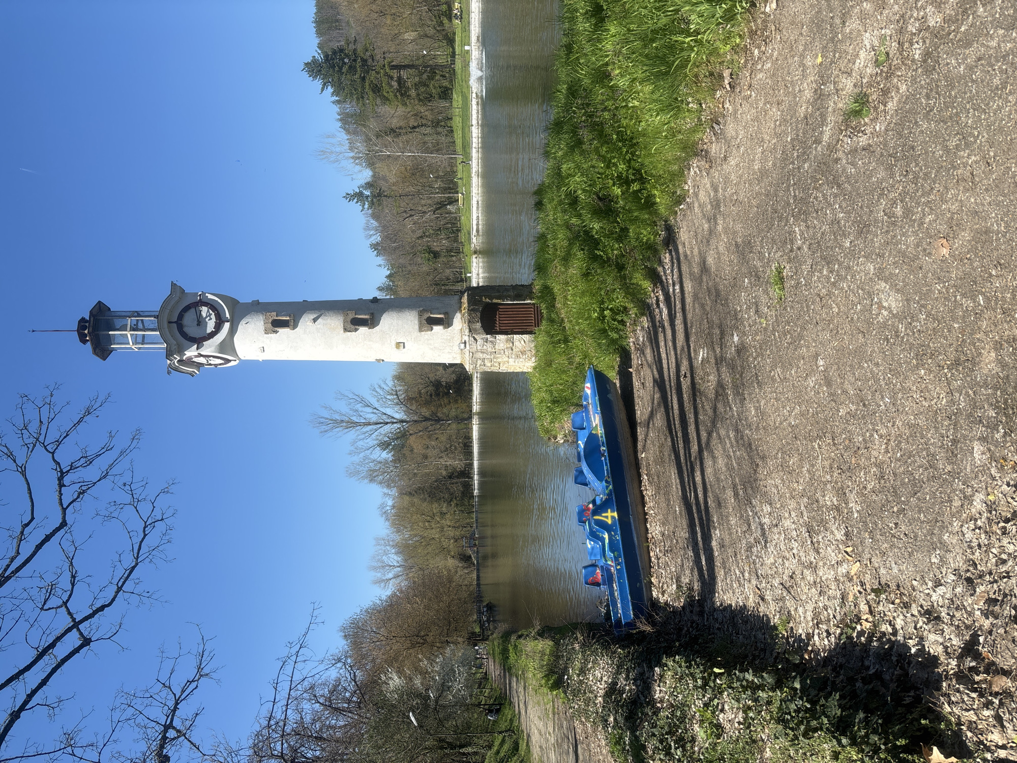 The image shows a bright sunny day in Pleven, Bulgaria, with the city's iconic clock tower prominently featured. The tower is a tall, white structure with a clock face near the top, positioned along a canal or lake. Alongside the canal, there is a paved path with green grass and vegetation. A blue pedal boat is resting on the side of the path, near the water's edge. Bare trees and evergreens are also visible in the background of the image.